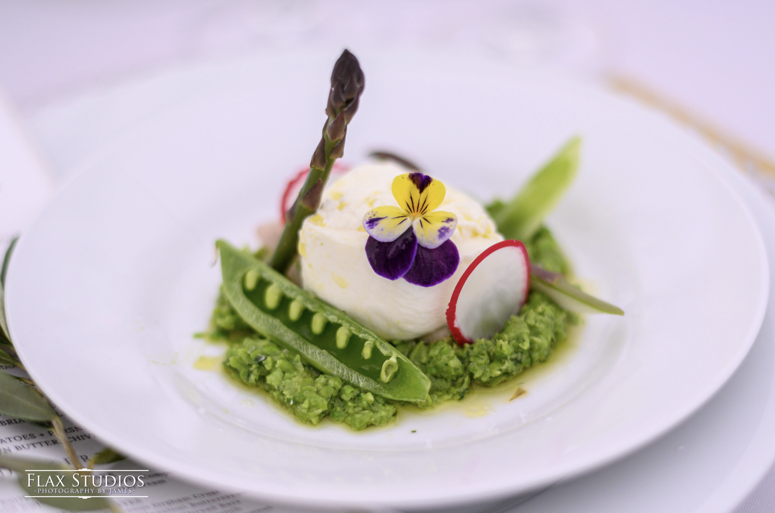 A gourmet dish featuring a scoop of white cheese or cream topped with edible purple and yellow flowers, surrounded by sliced radishes, snow peas, and a green vegetable puree or mash, served on a white plate.