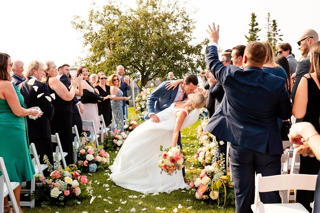 A wedding ceremony with a couple kissing while surrounded by guests. The bride is in a white gown, and the groom is in a blue suit. Guests are applauding and celebrating, with some raising their hands, outdoors with trees and floral decorations.