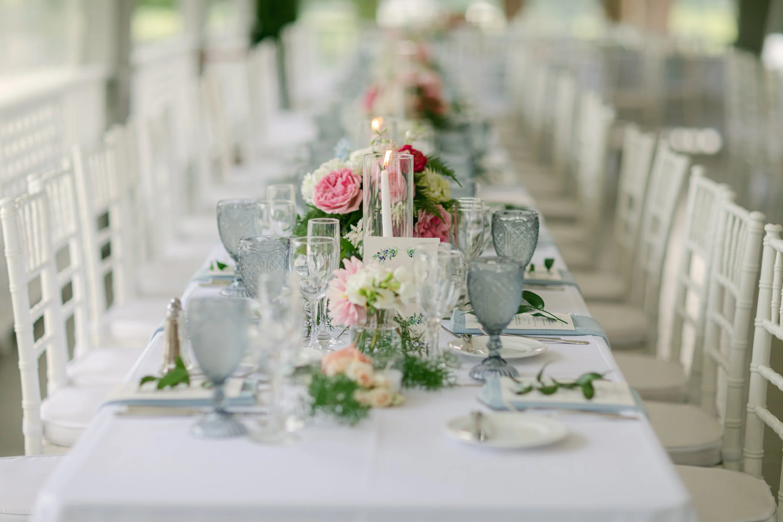 A long elegantly set dining table decorated with pink and white flowers, candles in glass holders, and multiple wine glasses, set for a formal event or wedding celebration.