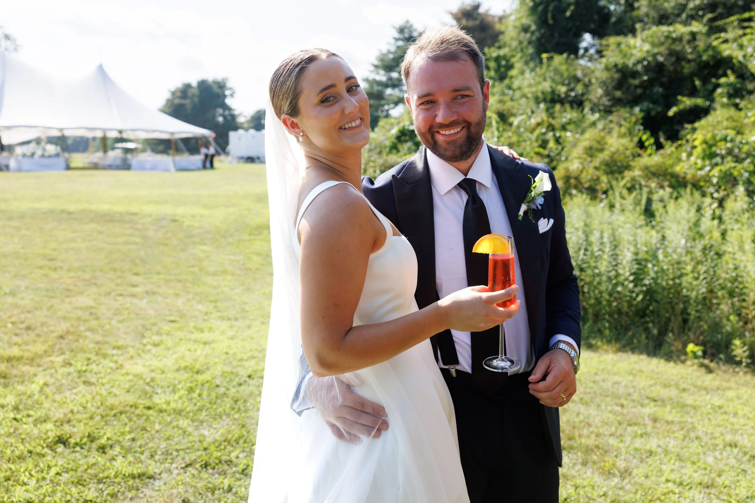 A happy bride and groom posing outdoors at a wedding reception, with the bride holding a glass of red-colored drink garnished with a lemon wedge, in a lush green field with tents and trees in the background.