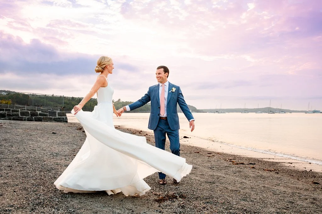 A bride and groom standing on a beach, holding hands and gazing at each other during sunset.