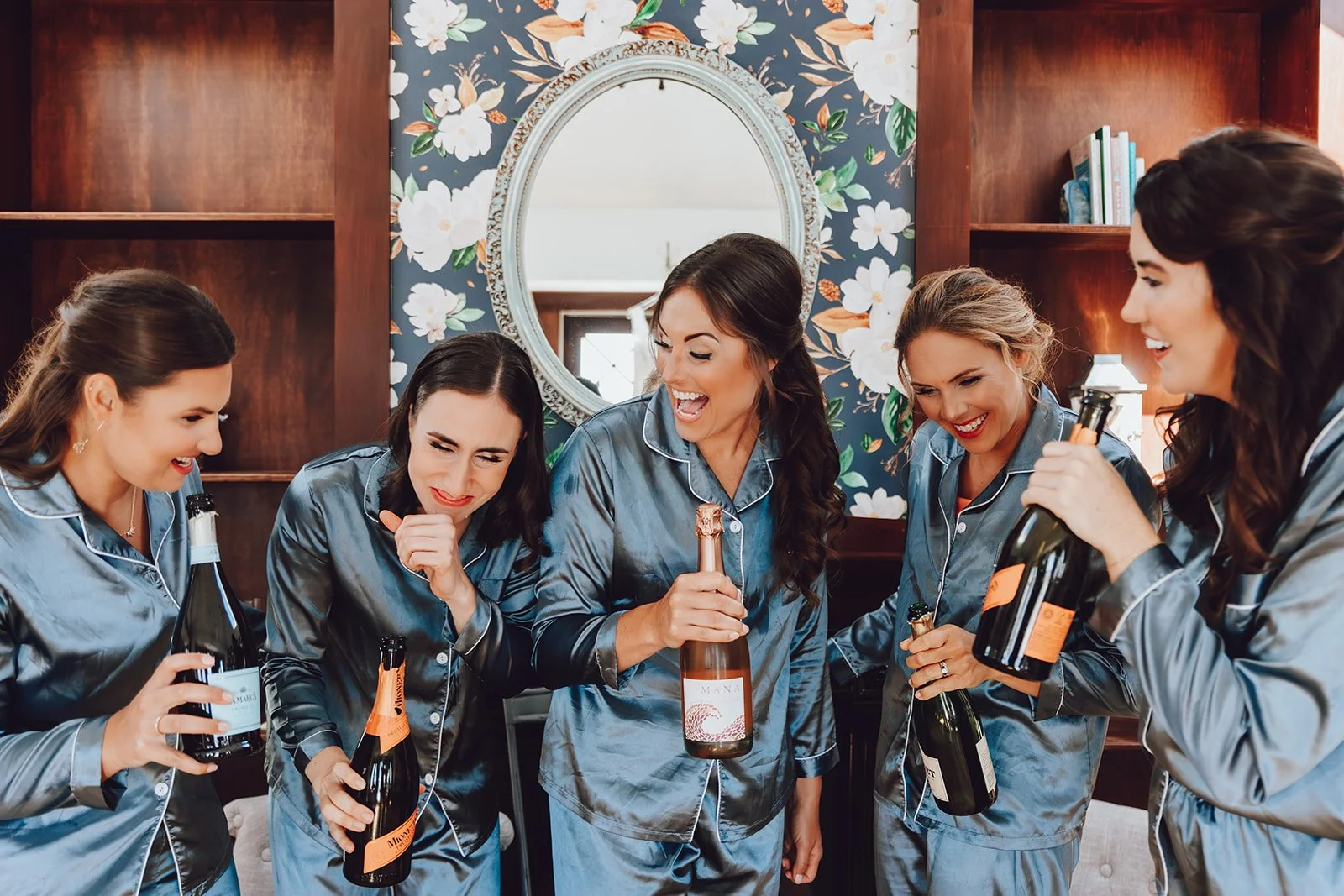 Group of five women in matching satin pajamas celebrating with bottles of champagne in a cozy, floral-decorated room.