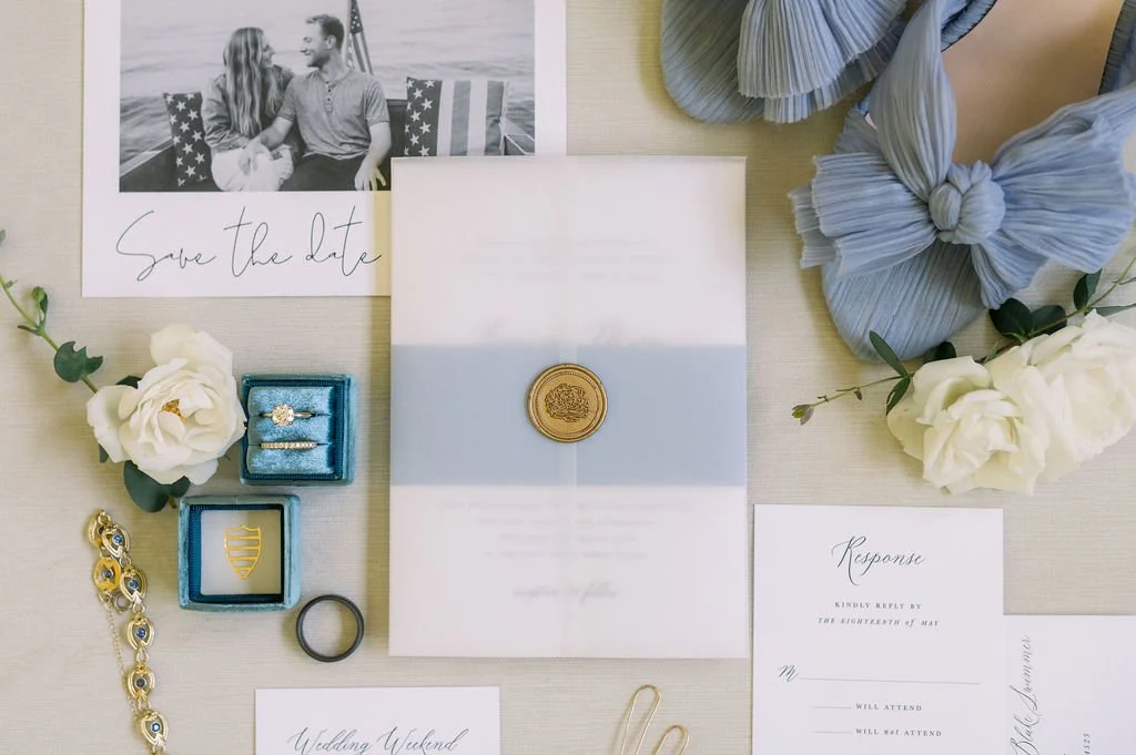 Wedding invitation with a gray ribbon seal, surrounded by a 'Save the Date' card, a response card, white flowers, jewelry including rings and a bracelet, and a blue fabric bow, all arranged on a light-colored surface.
