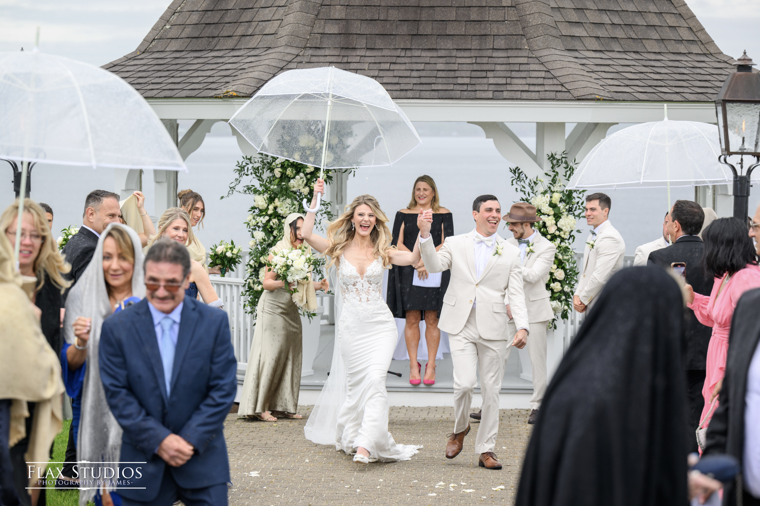 Bride and groom walking hand in hand under umbrellas, smiling, surrounded by wedding guests at an outdoor wedding ceremony in rainy weather.