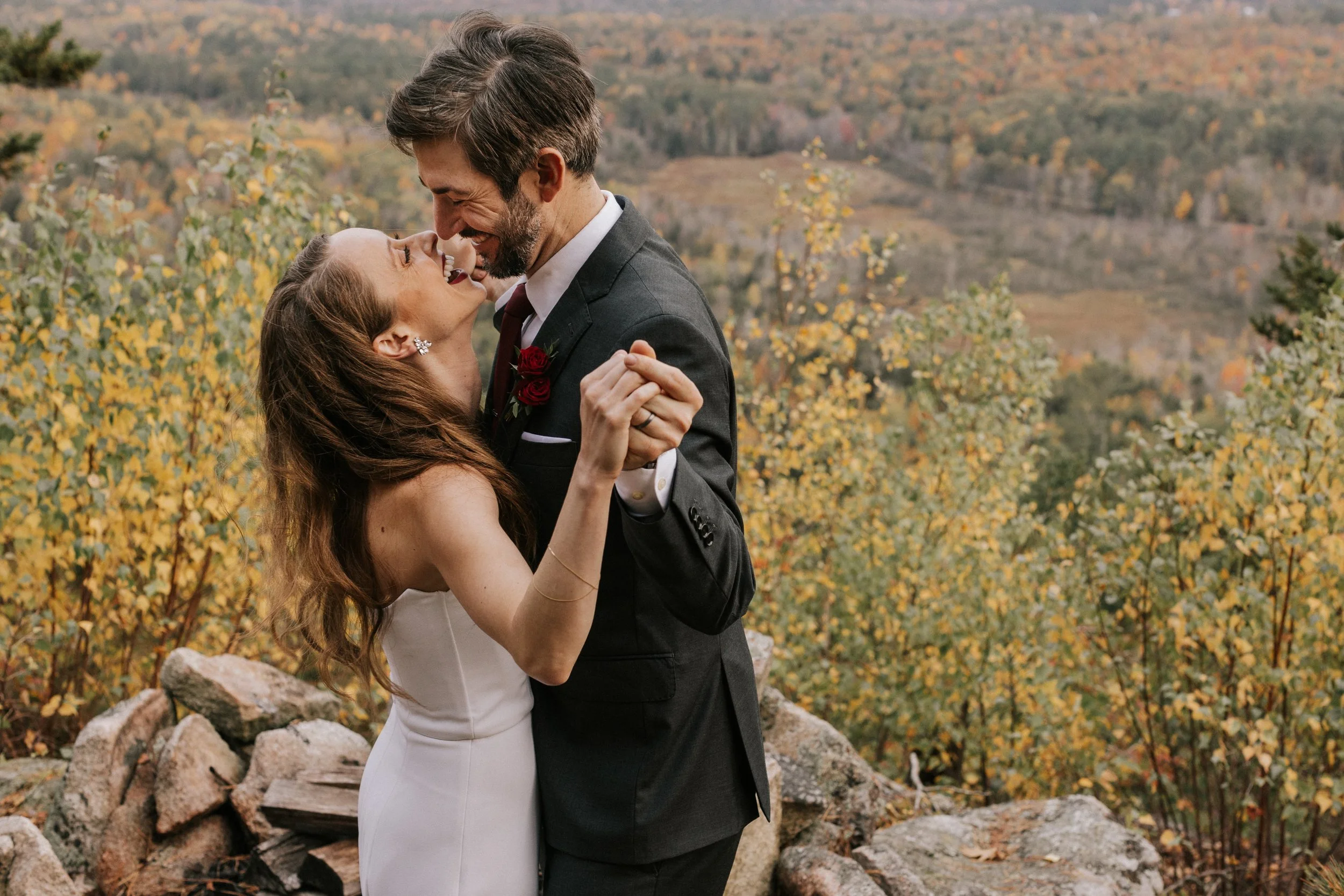 A newlywed couple dancing outdoors on a rocky terrain surrounded by autumn foliage with trees in yellow, orange, and green, and hills in the background.