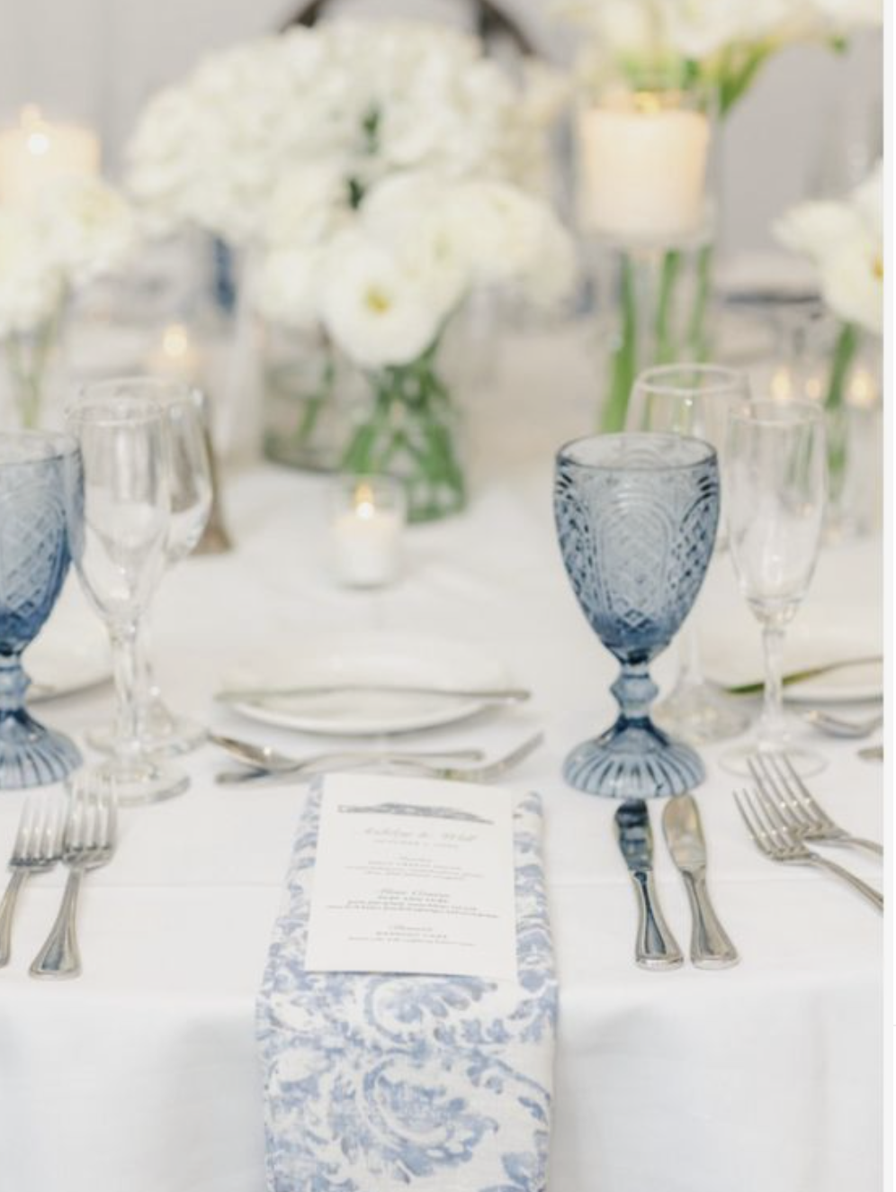 Elegant table setting with blue and clear glassware, silverware, a floral menu, and a centerpiece of white flowers and candles.