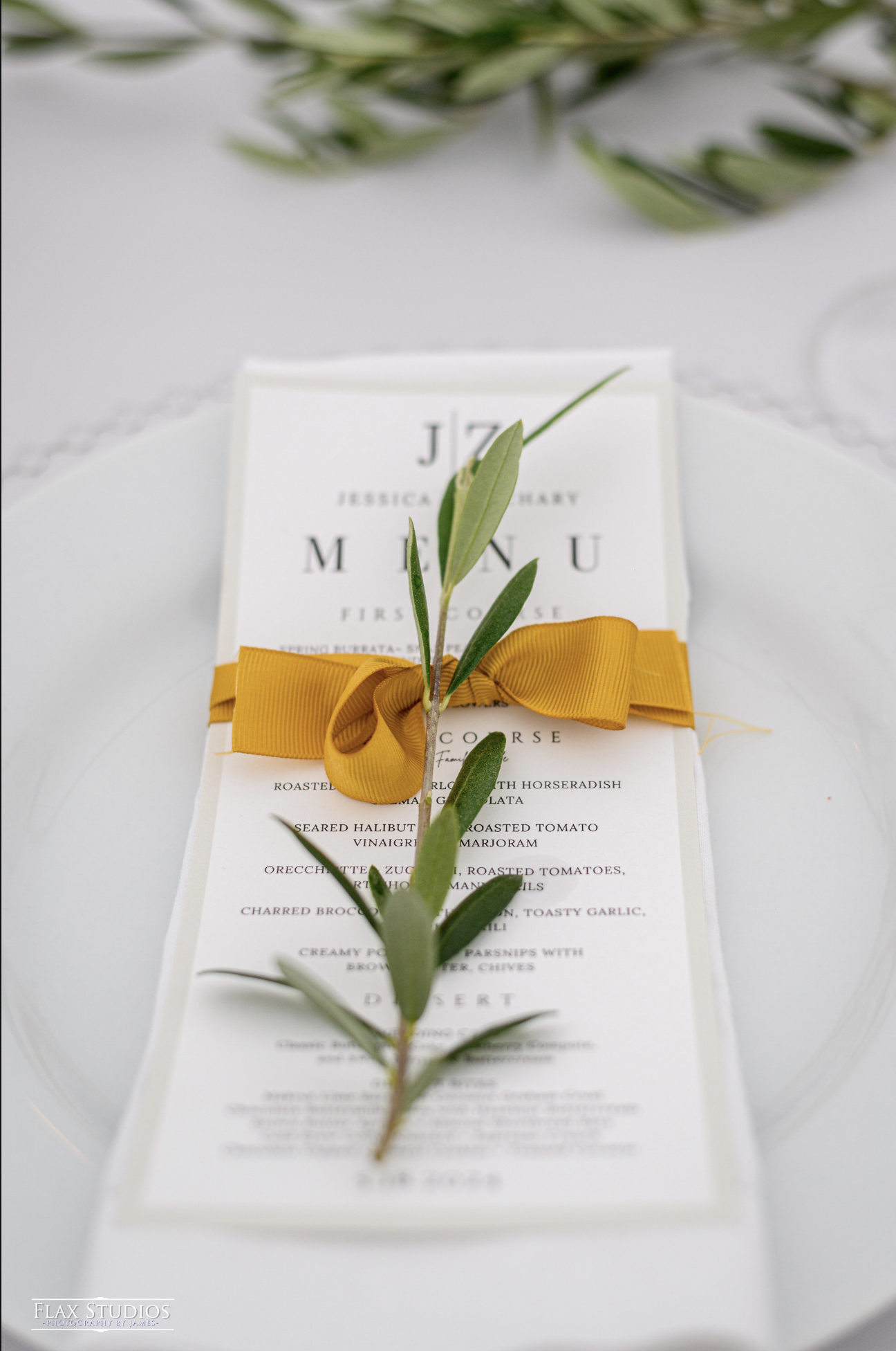 A wedding menu card decorated with an olive branch and yellow ribbon, placed on a white plate.