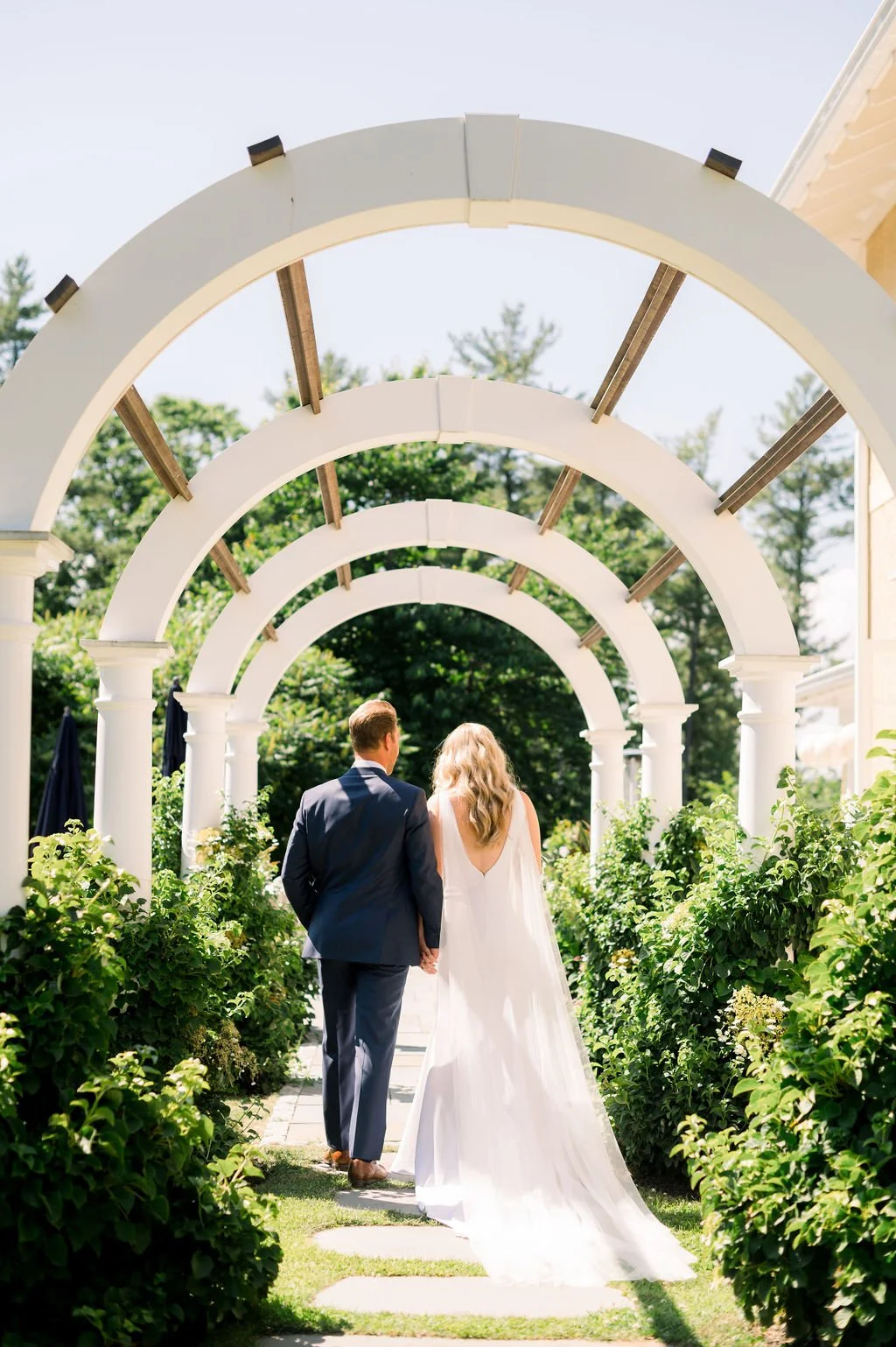 A bride and groom walking hand in hand through a garden archway on a sunny day, with greenery and trees in the background.