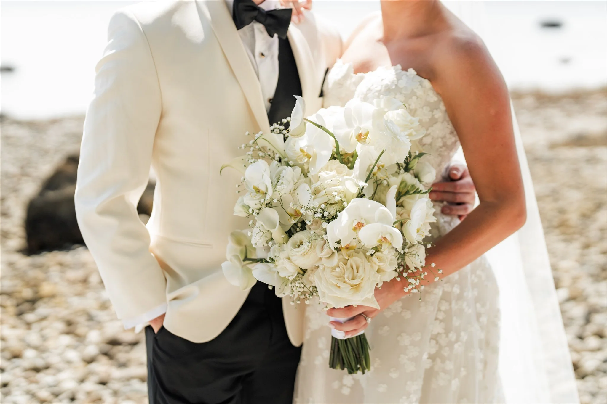 A bride and groom on their wedding day, with the bride holding a large bouquet of white flowers, including orchids and roses, on a beach.