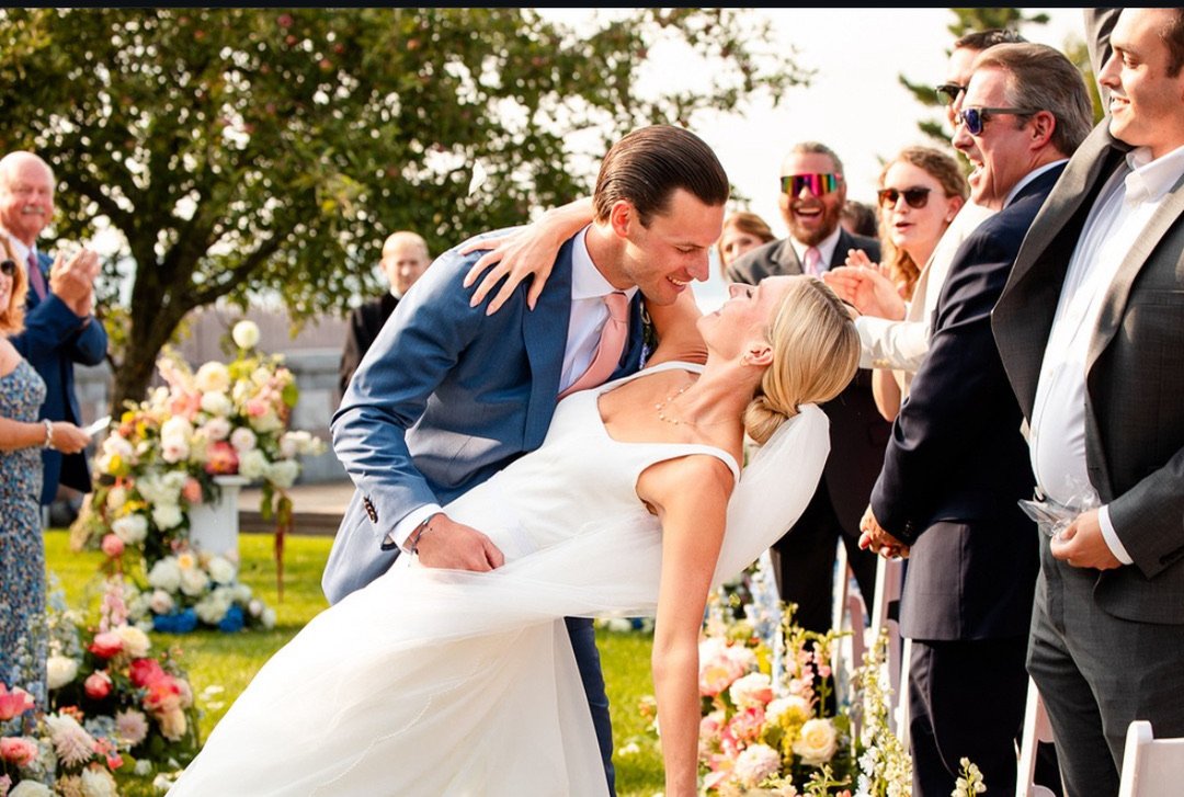 A bride and groom sharing a dance outdoors during their wedding ceremony with guests clapping and smiling around them, lush green trees in the background.