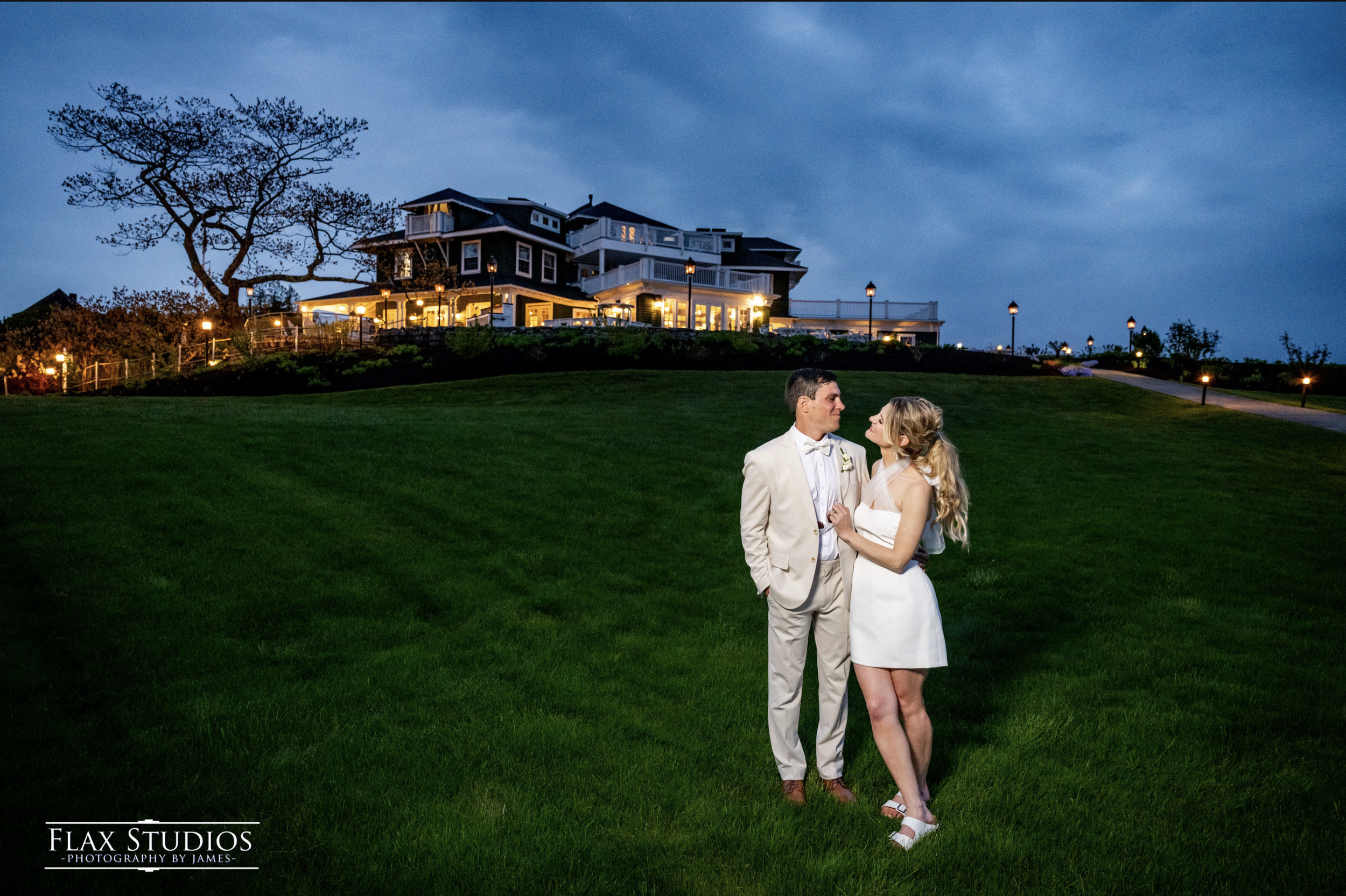 Couple in wedding attire standing on a grassy hilltop at dusk, with a large house illuminated in the background.