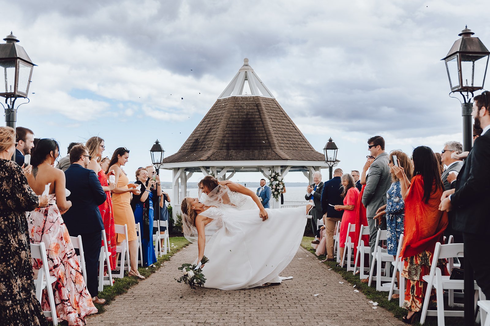 A bride and groom celebrate at an outdoor wedding ceremony near a gazebo, with guests on either side taking photos and videos. The bride is holding a bouquet and is leaning forward as the groom spins or dips her against a cloudy sky backdrop.
