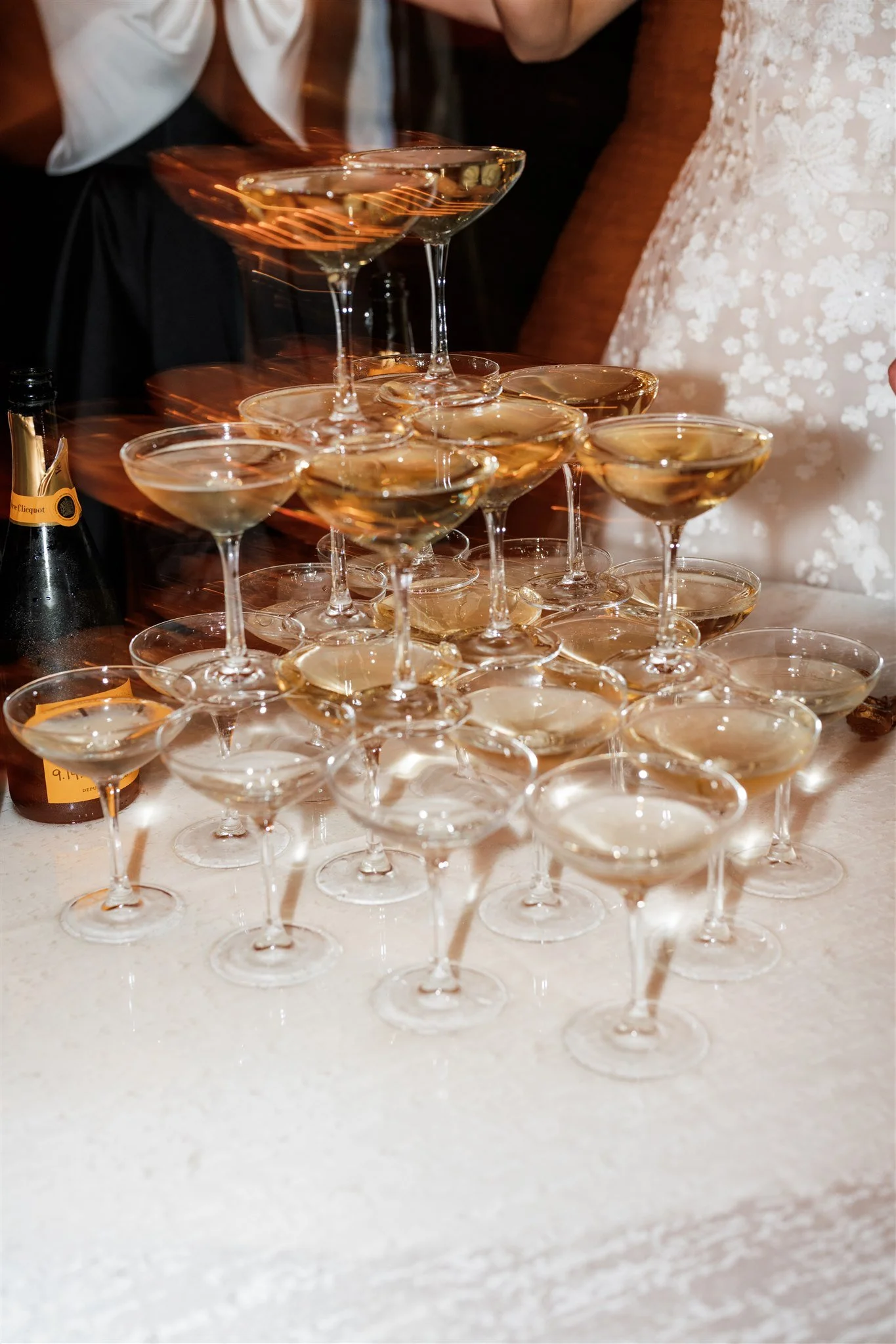 A pyramid of champagne glasses filled with champagne on a white table, with people dressed formally in the background.
