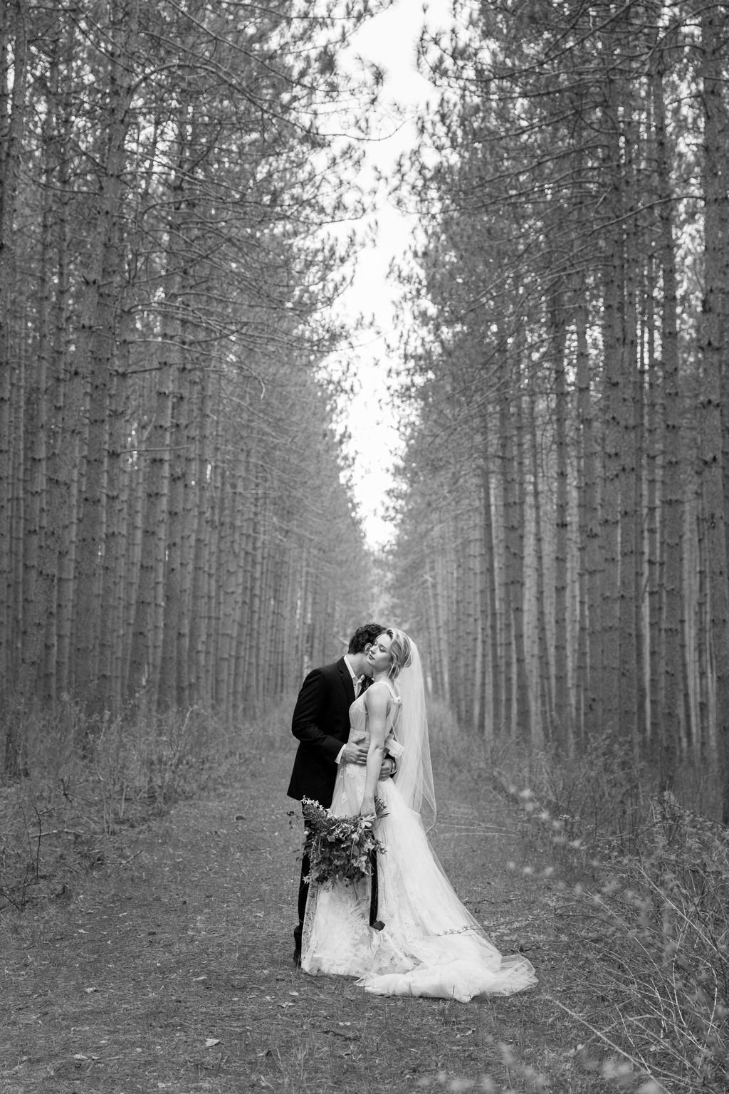 A black and white photo of a bride and groom sharing a kiss in a forest, with tall trees lining the path.