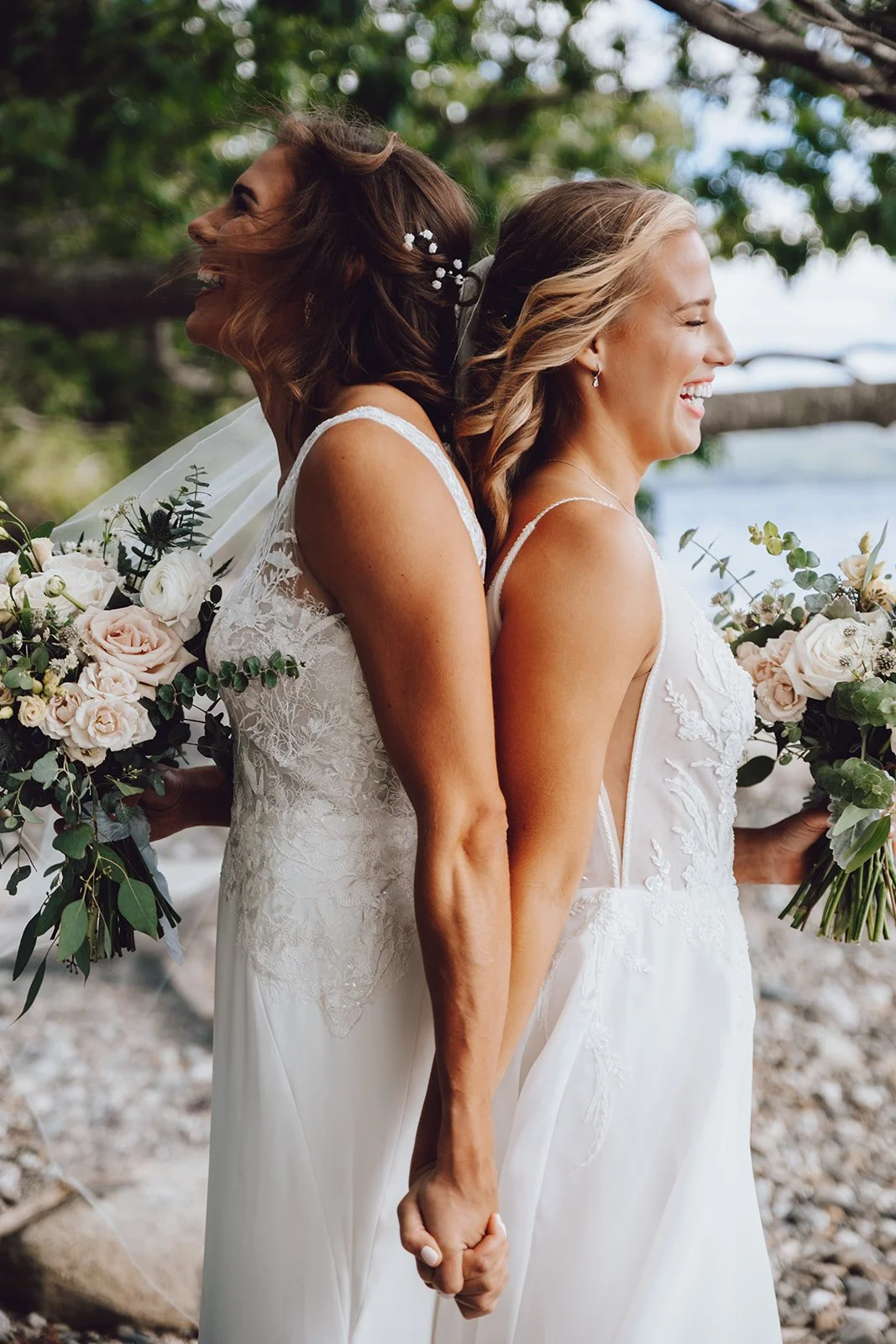 Two women in white wedding dresses are back to back, holding hands, smiling, and holding bouquets of flowers, outdoors with trees and water in the background.