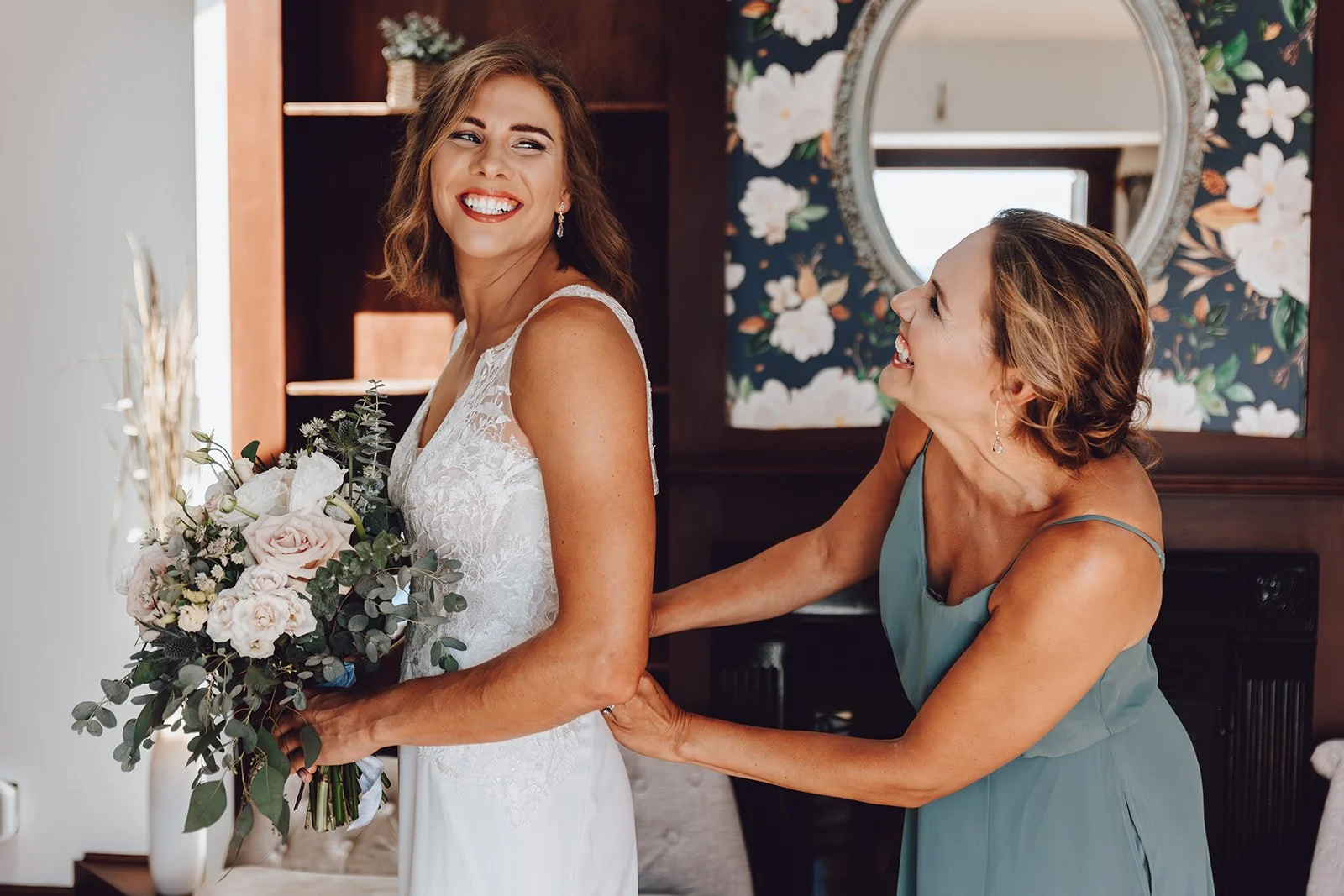 A bride in a white lace wedding dress holding a bouquet of flowers, smiling while being supported from behind by a woman in a teal dress, in a room with floral wallpaper and a mirror.