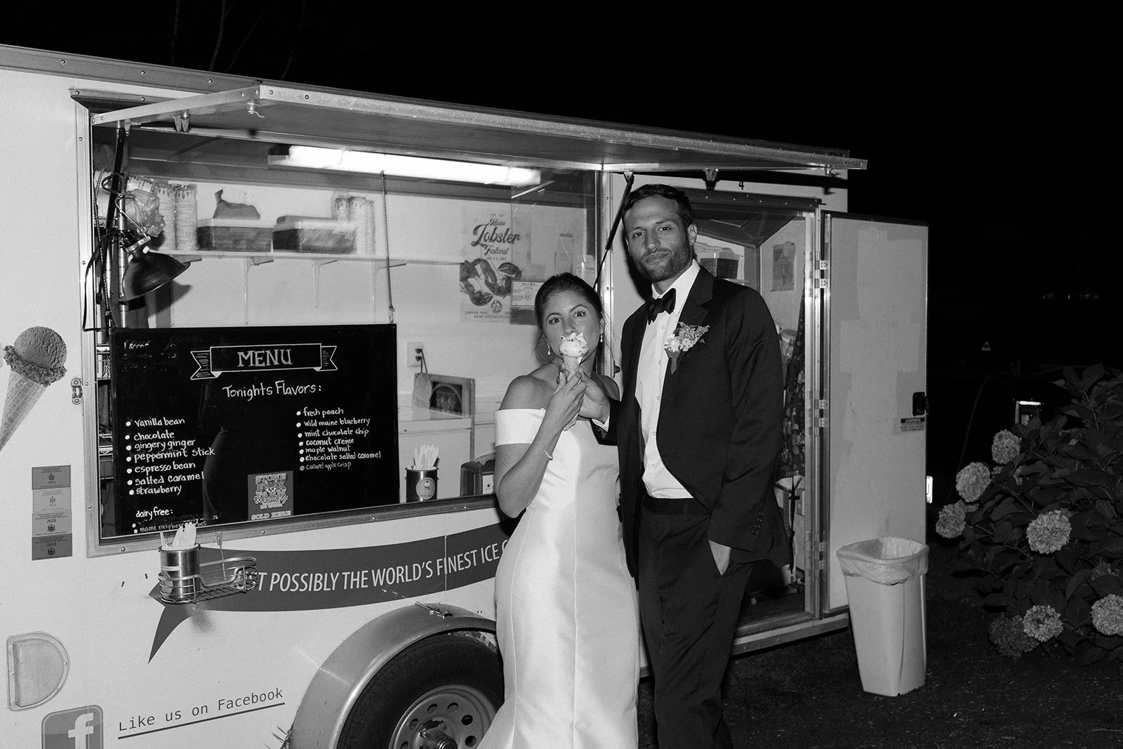 Black and white photo of a newlywed couple in wedding attire standing in front of an ice cream truck at night. The bride is holding an ice cream cone, and the groom has one hand in his pocket, smiling.