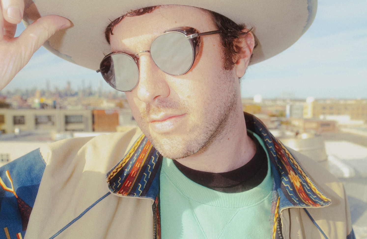 Close-up of a man wearing a hat, round sunglasses, and a colorful patterned jacket, standing outdoors with a cityscape in the background.