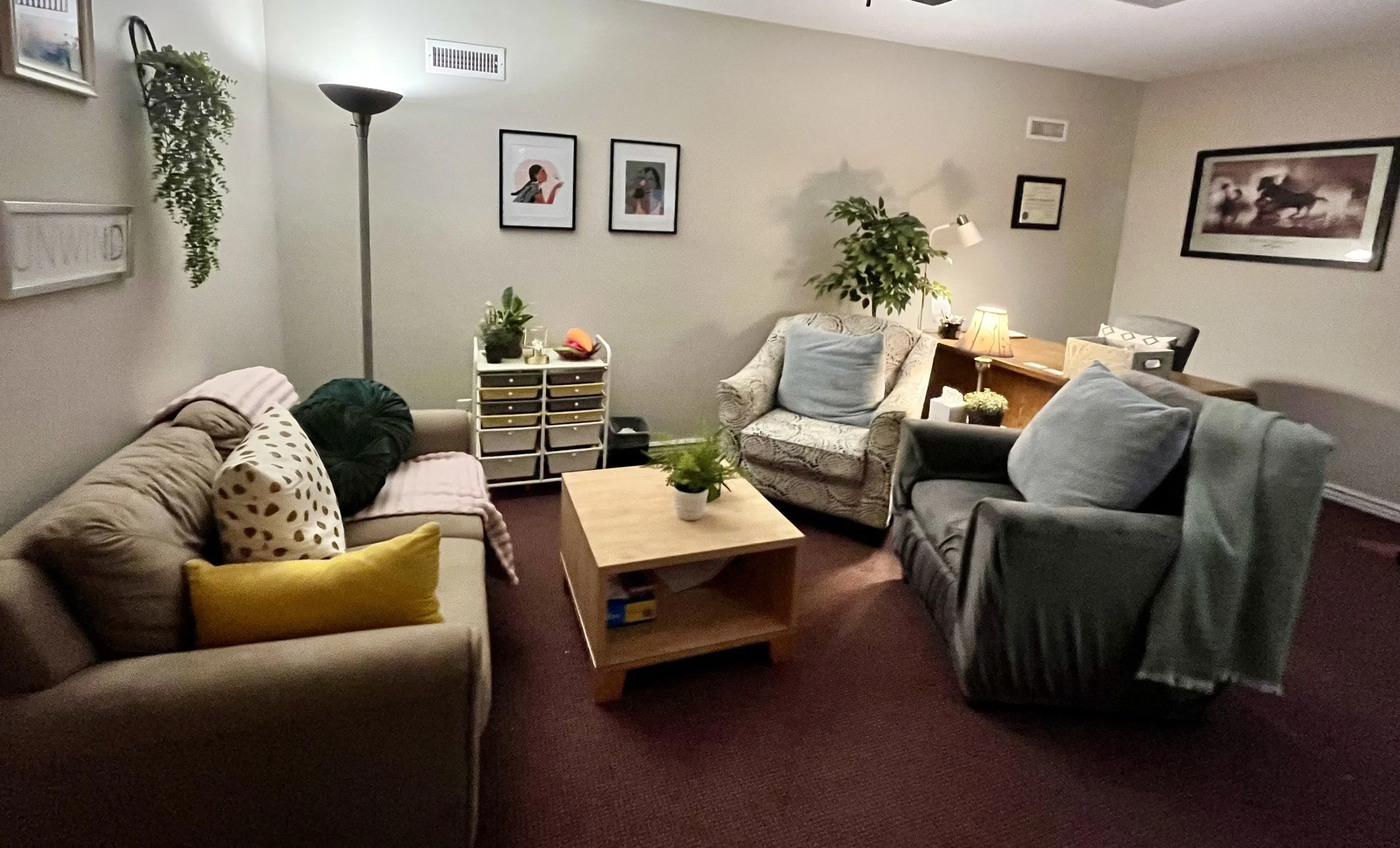 Living room with beige and patterned chairs, wooden coffee table with snacks and tissue box, wall art of two women, small storage cart with water bottles and cleaning supplies, floor lamp, small fan, and carpet.