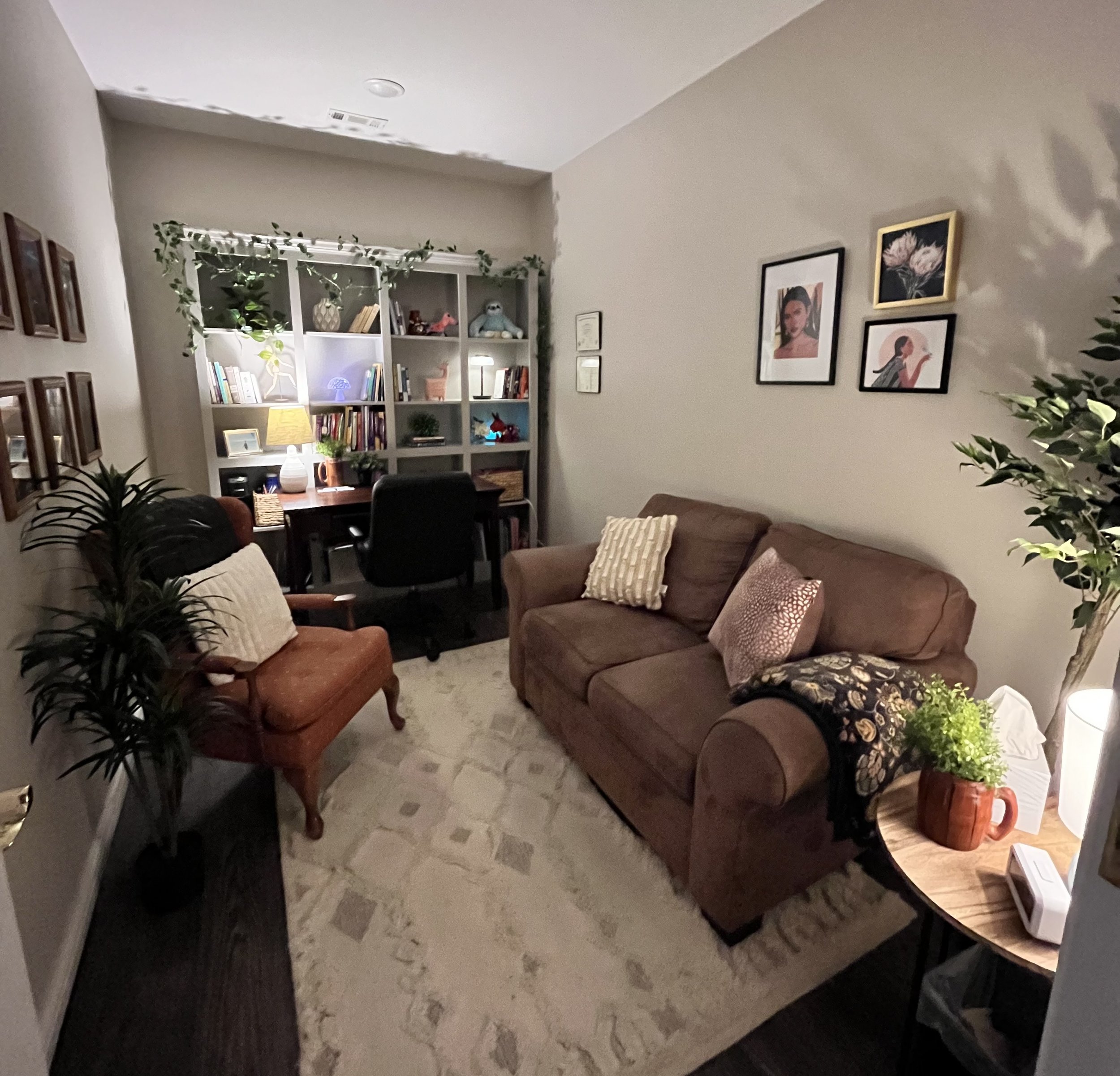 A cozy living room with a beige patterned rug, a brown sofa with a pink pillow, a smaller chocolate-colored armchair with a white pillow, a small side table with decorations, a dark wooden desk with a black office chair, and a large gray bookshelf filled with books and decorative items.
