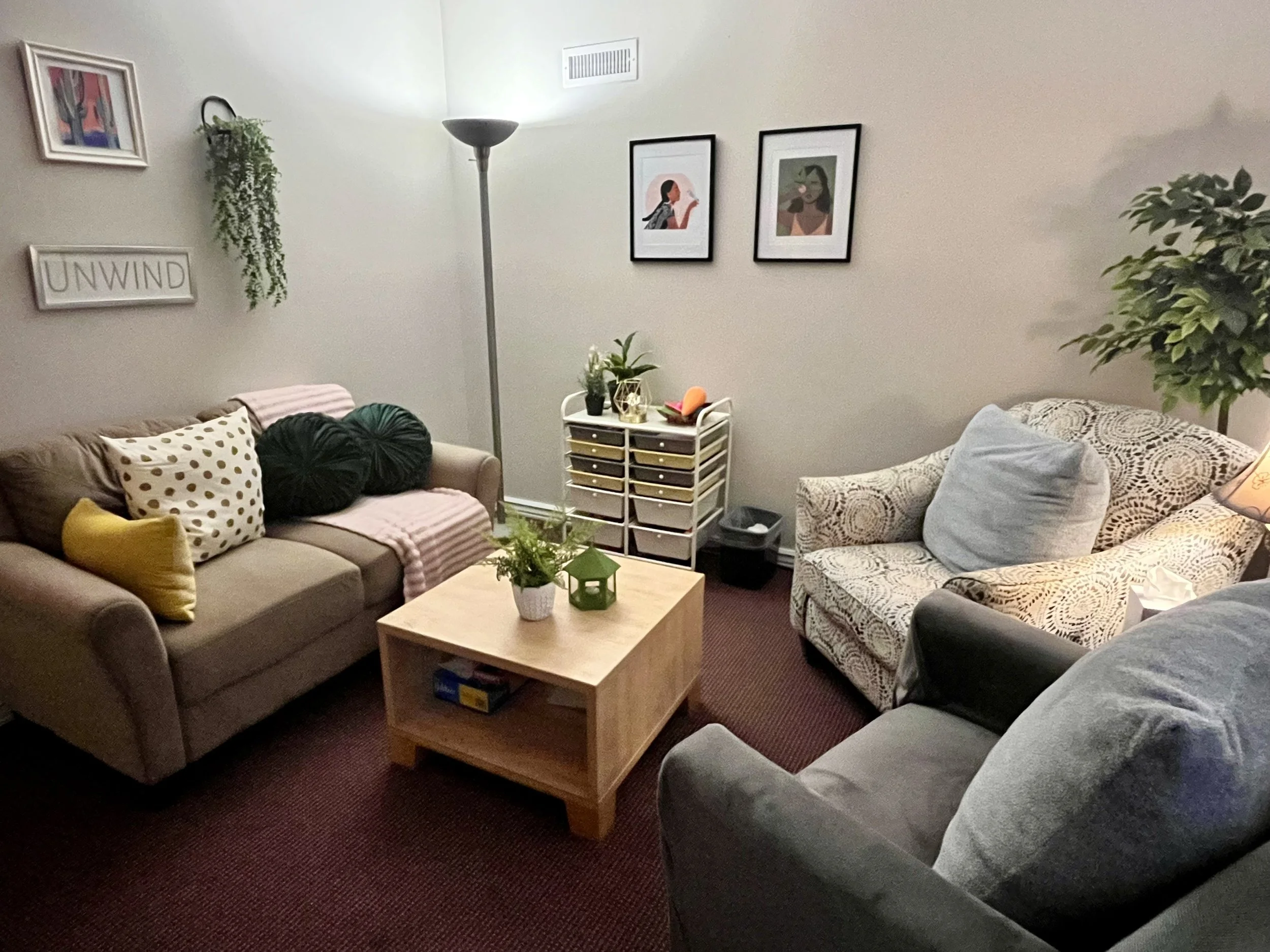 A beige sofa with three decorative pillows on it, a throw blanket, a floor lamp next to the sofa, a small fan on the floor, a storage cart with various items, and framed art and a sign that says 'UNWIND' on the wall.
