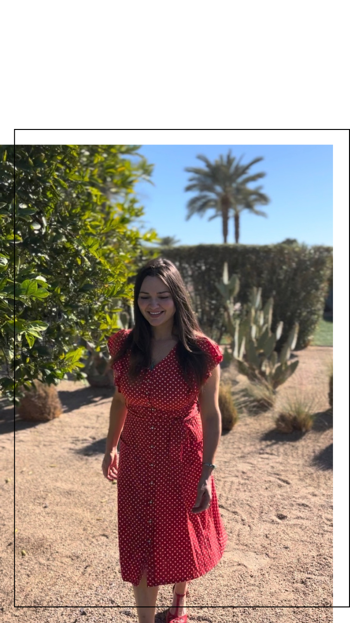 A woman in a red dress with white polka dots walking outdoors on a sunny day, surrounded by desert plants, including cacti and palm trees.