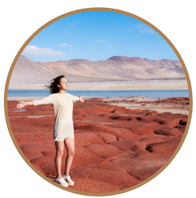 A woman standing with arms outstretched on red rocks by a lake, with mountains in the background under a blue sky.