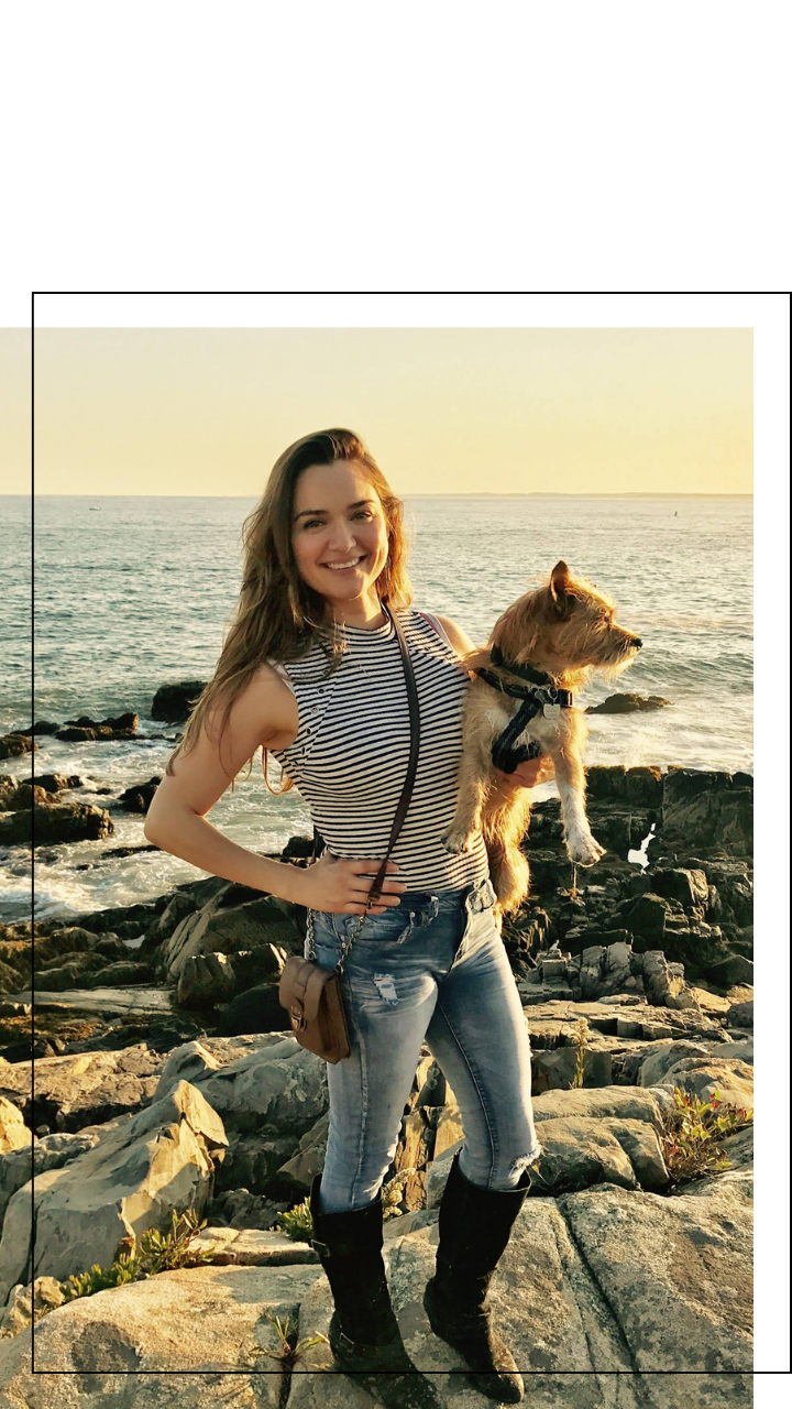 A woman with long hair wearing a striped sleeveless top, ripped jeans, and black boots, holding a small dog in her arms by the rocky shoreline during sunset.