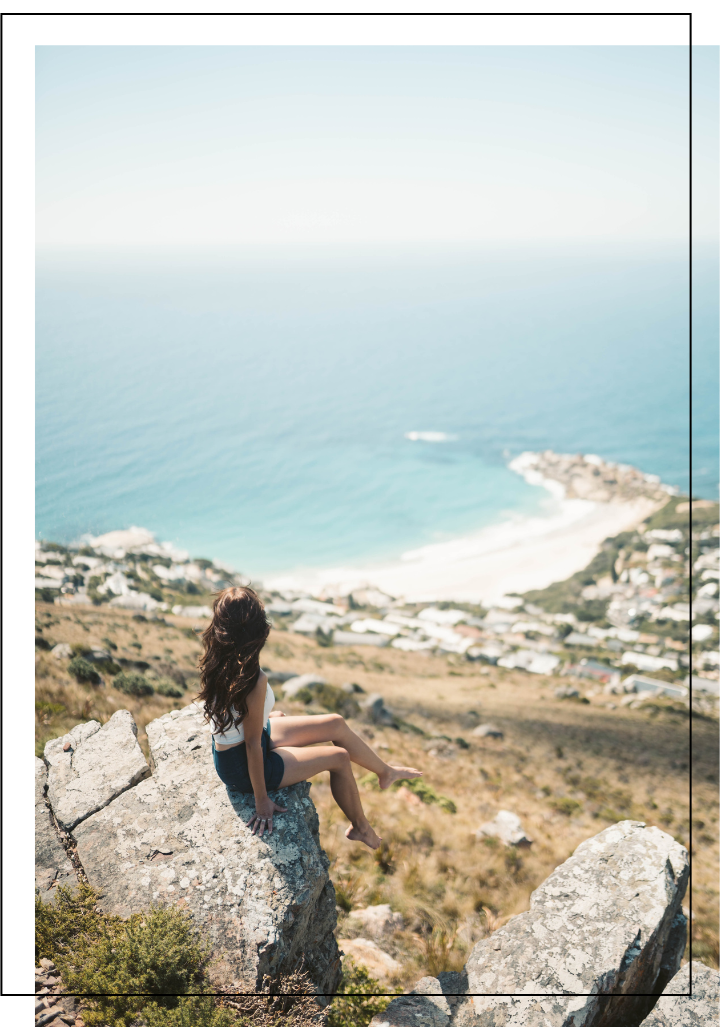 Young woman with long brown hair sitting on a large rock overlooking a coastal landscape with a beach, houses, and the ocean.