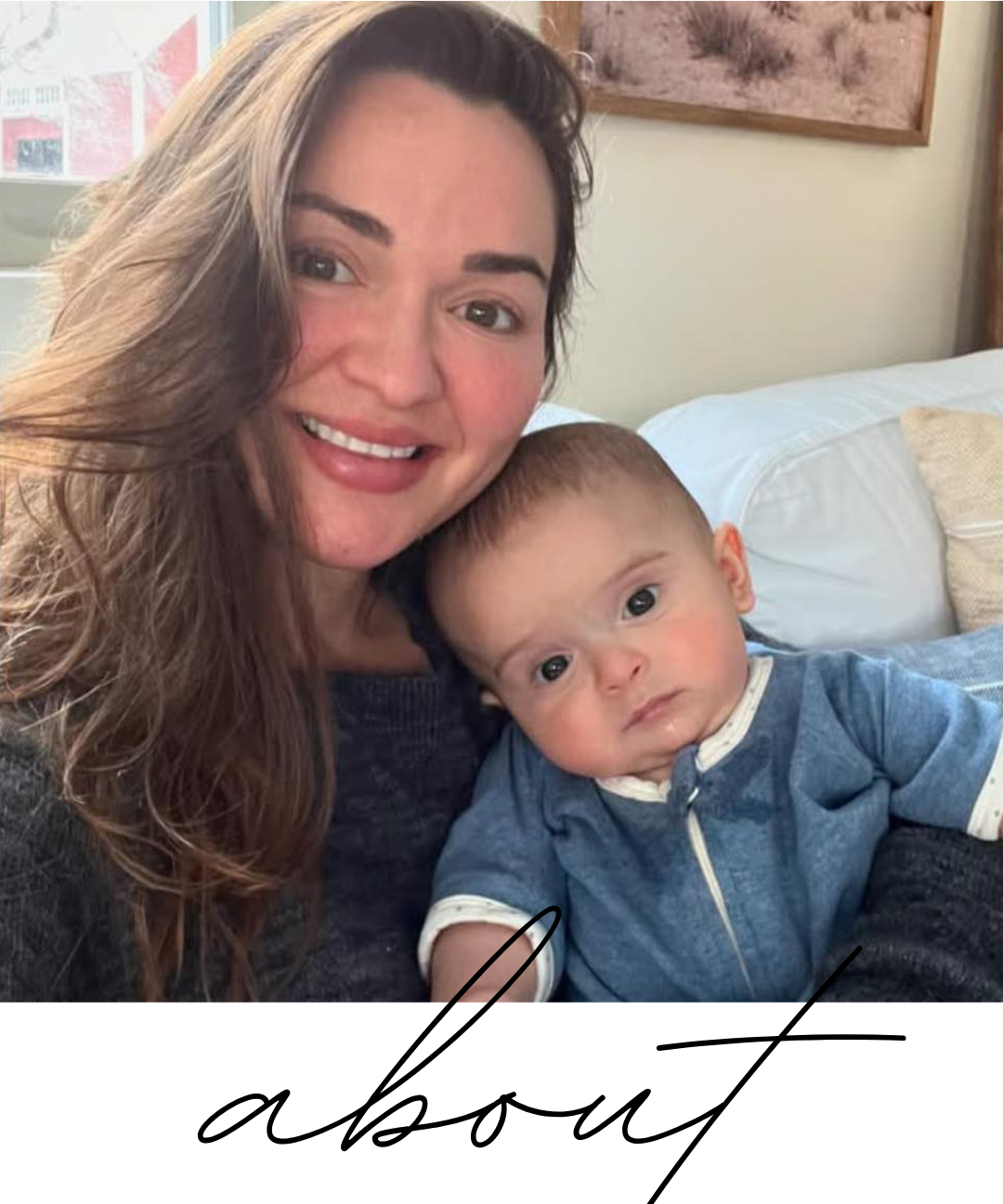 A woman with long brown hair smiling and holding a baby with short light brown hair, both sitting on a bed in a room with a framed picture on the wall behind them.