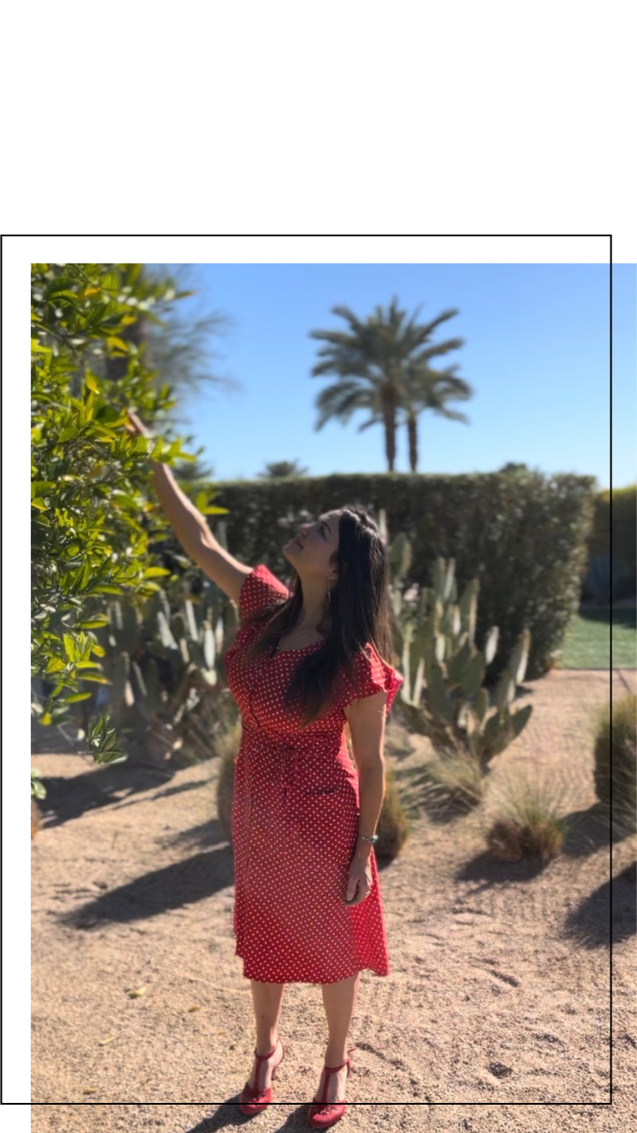 A woman in a red polka dot dress and red shoes standing outdoors in a desert-like garden, reaching up to touch the leaves of a plant, with palm trees and blue sky in the background.