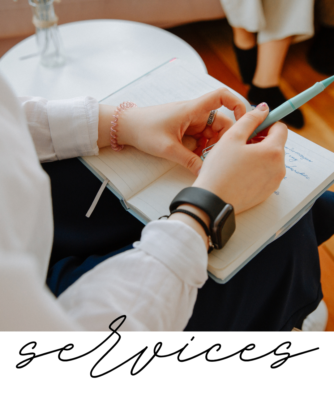 Person sitting and writing in a notebook with a pen, notes visible on the page, wearing a smartwatch and a pink hair tie, with another person sitting nearby in the background.
