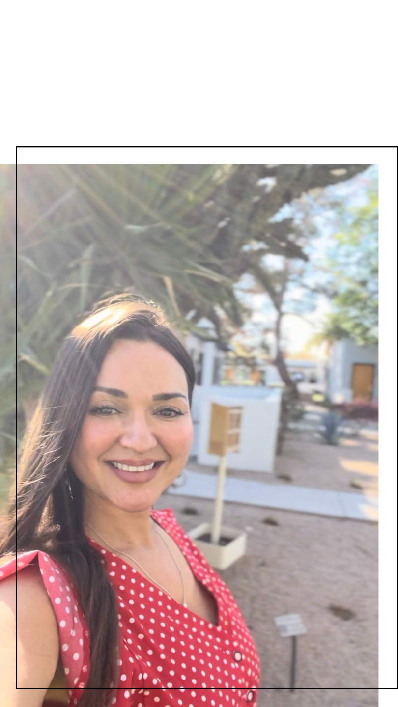 A woman with long dark hair and light makeup takes a selfie outdoors, smiling at the camera. She is wearing a red polka dot dress and stands in front of outdoor structures and trees, with sunlight and a clear sky in the background.