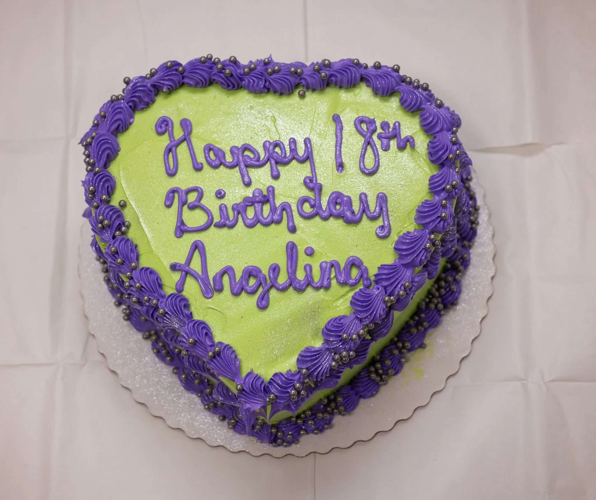 Heart-shaped cake with "Happy 18th Birthday Angelina" in purple icing on neon green frosting, decorated with purple icing border and silver sprinkles, placed on a white surface.