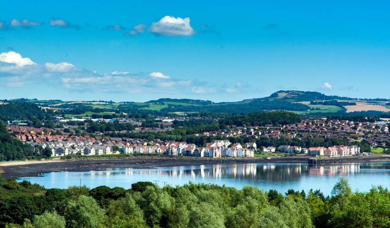 A scenic view of Dalgety Bay, with colourful houses and rolling green hills in the background under a partly cloudy blue sky.