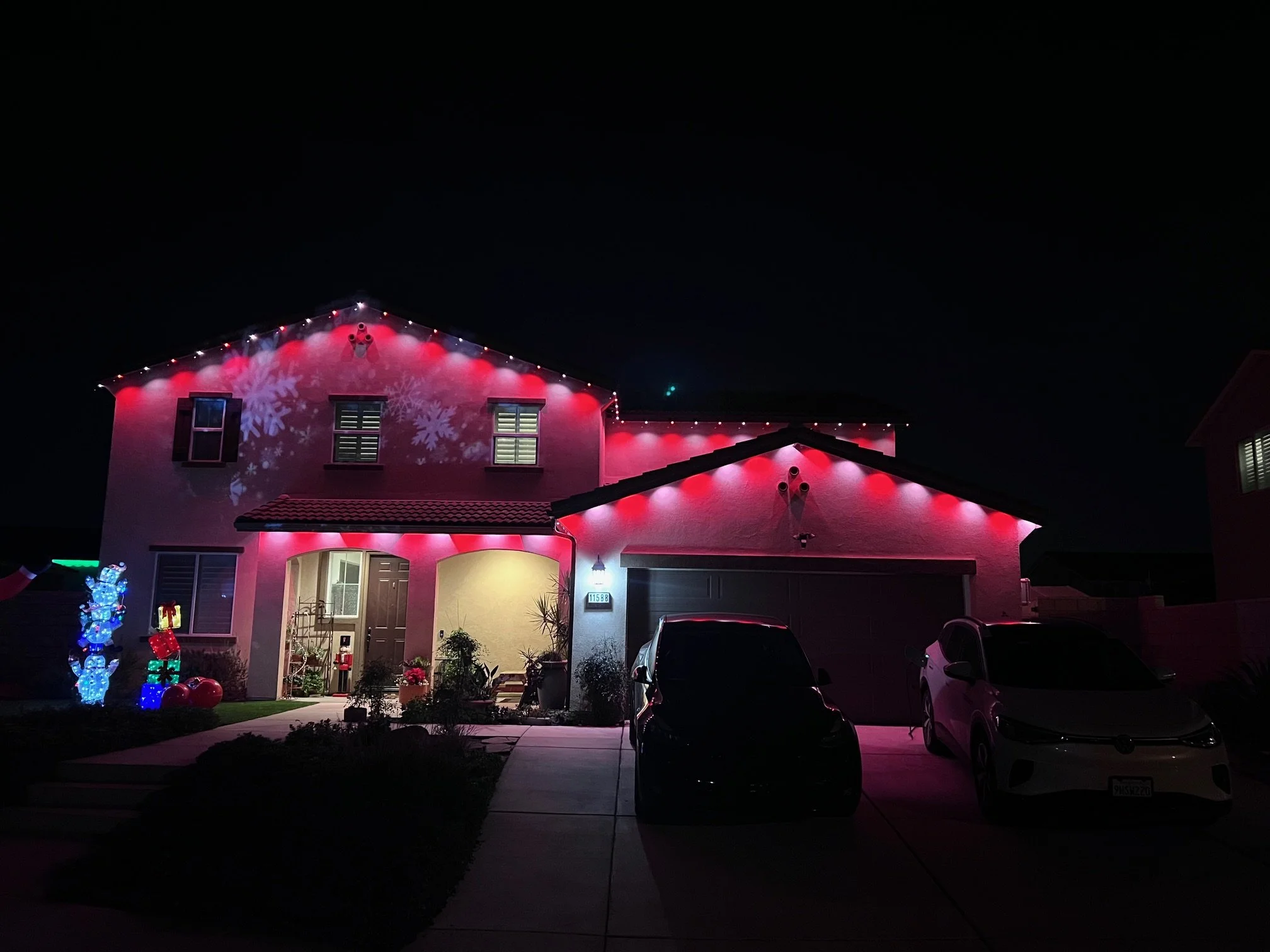 A two-story house decorated with red and white holiday lights and snowflake projections at night. The front yard has illuminated holiday figures, and the driveway has two cars parked outside.