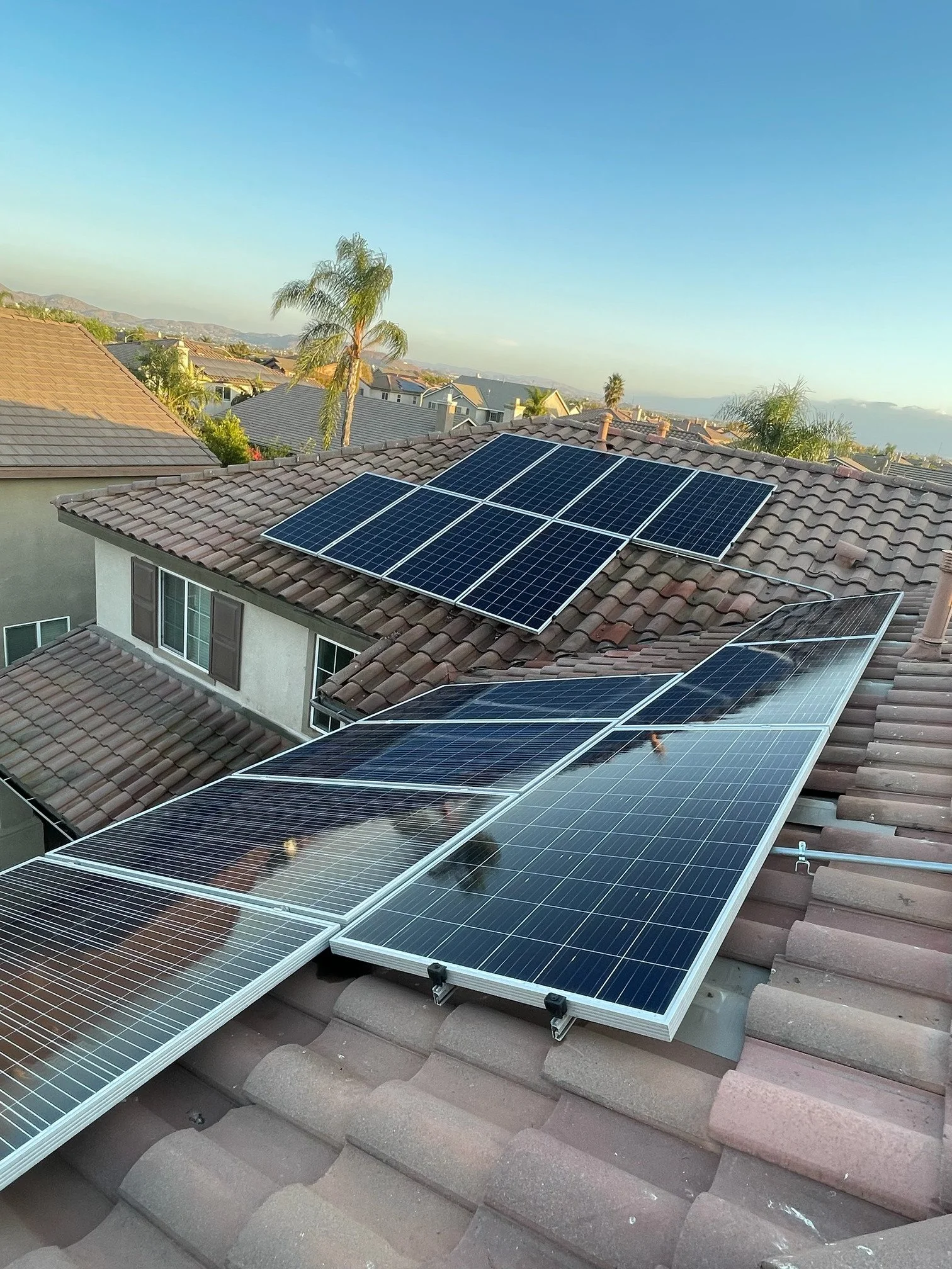 Multiple solar panels installed on a brown tiled roof in a suburban neighborhood, with trees and houses in the background under a clear sky.
