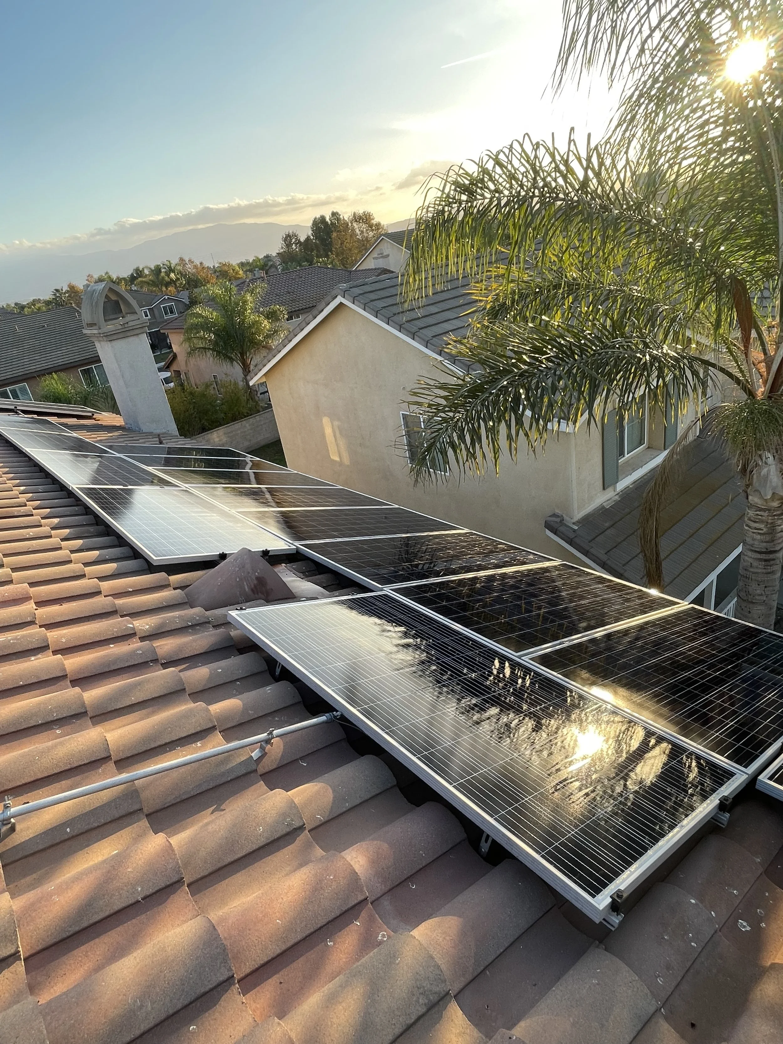 Sunset over a residential neighborhood with solar panels installed on a tiled roof, palm trees, and neighboring houses in the background.