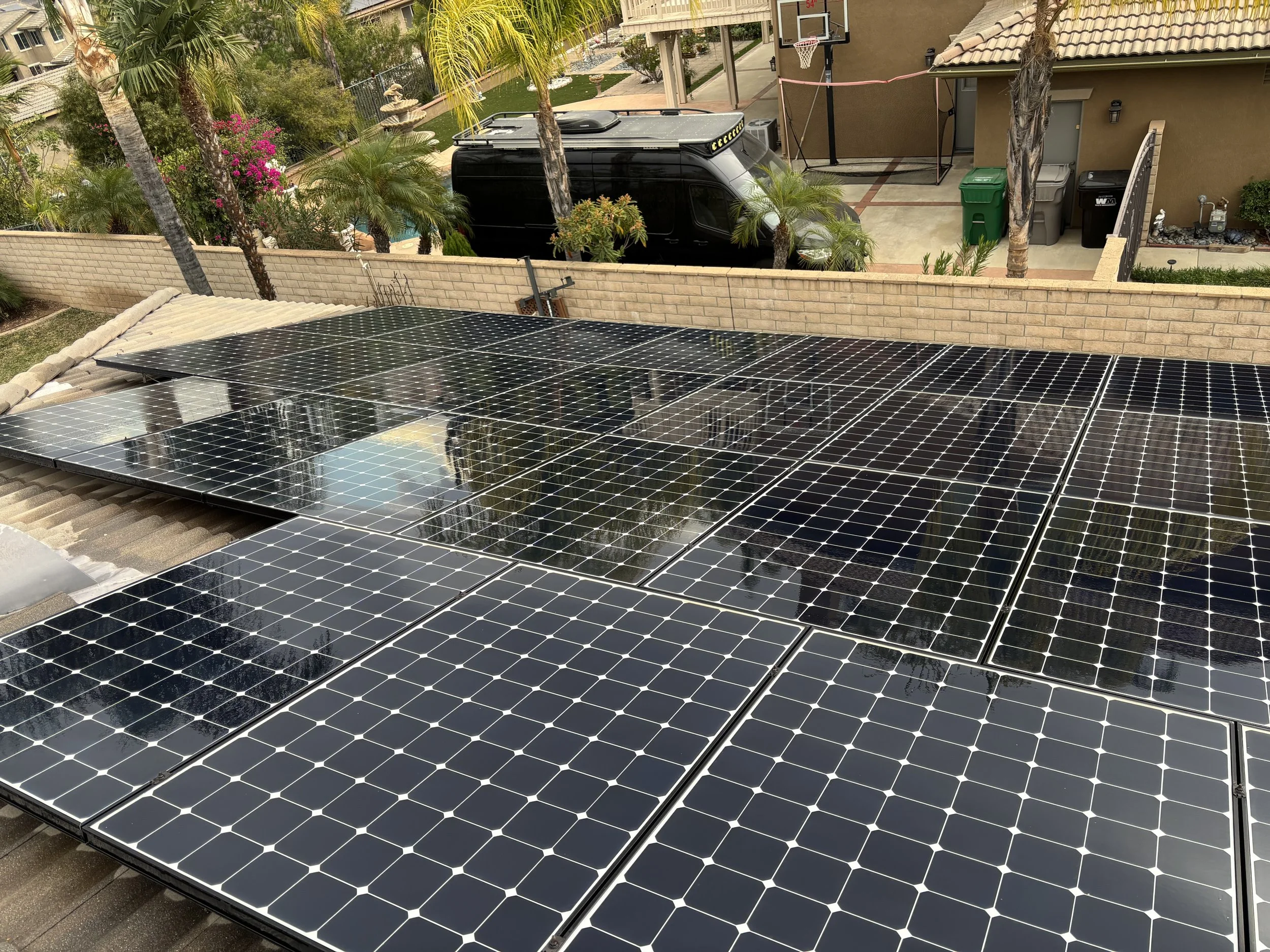 Multiple black solar panels installed on a residential roof with palm trees, a brick wall, and a backyard with a basketball hoop in the background.