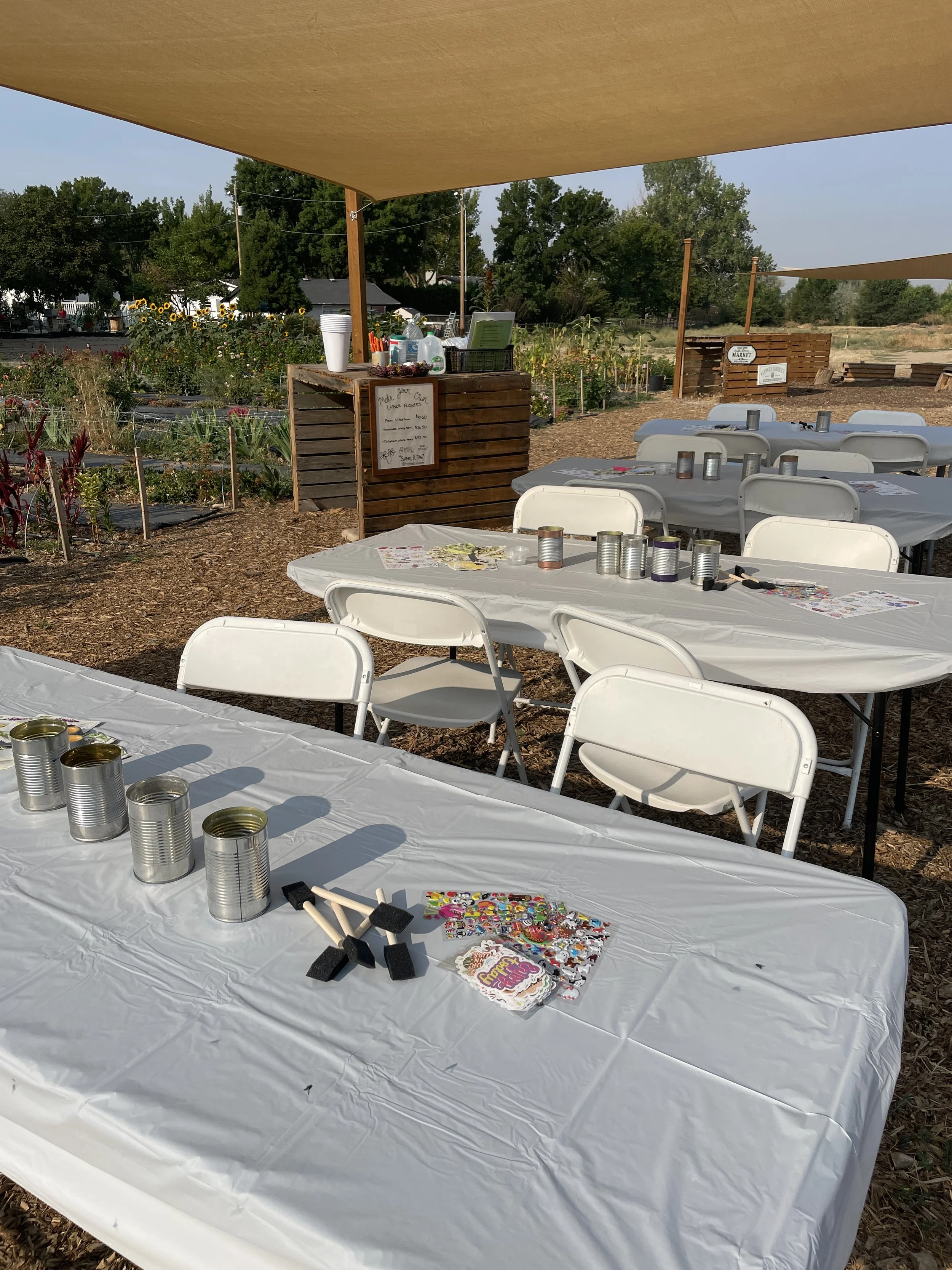 Outdoor event setup with tables covered in white tablecloths, decorated with tins, foam stamps, and craft supplies, in a farm setting with sunflowers and plants, shaded by large beige canopy sheets.