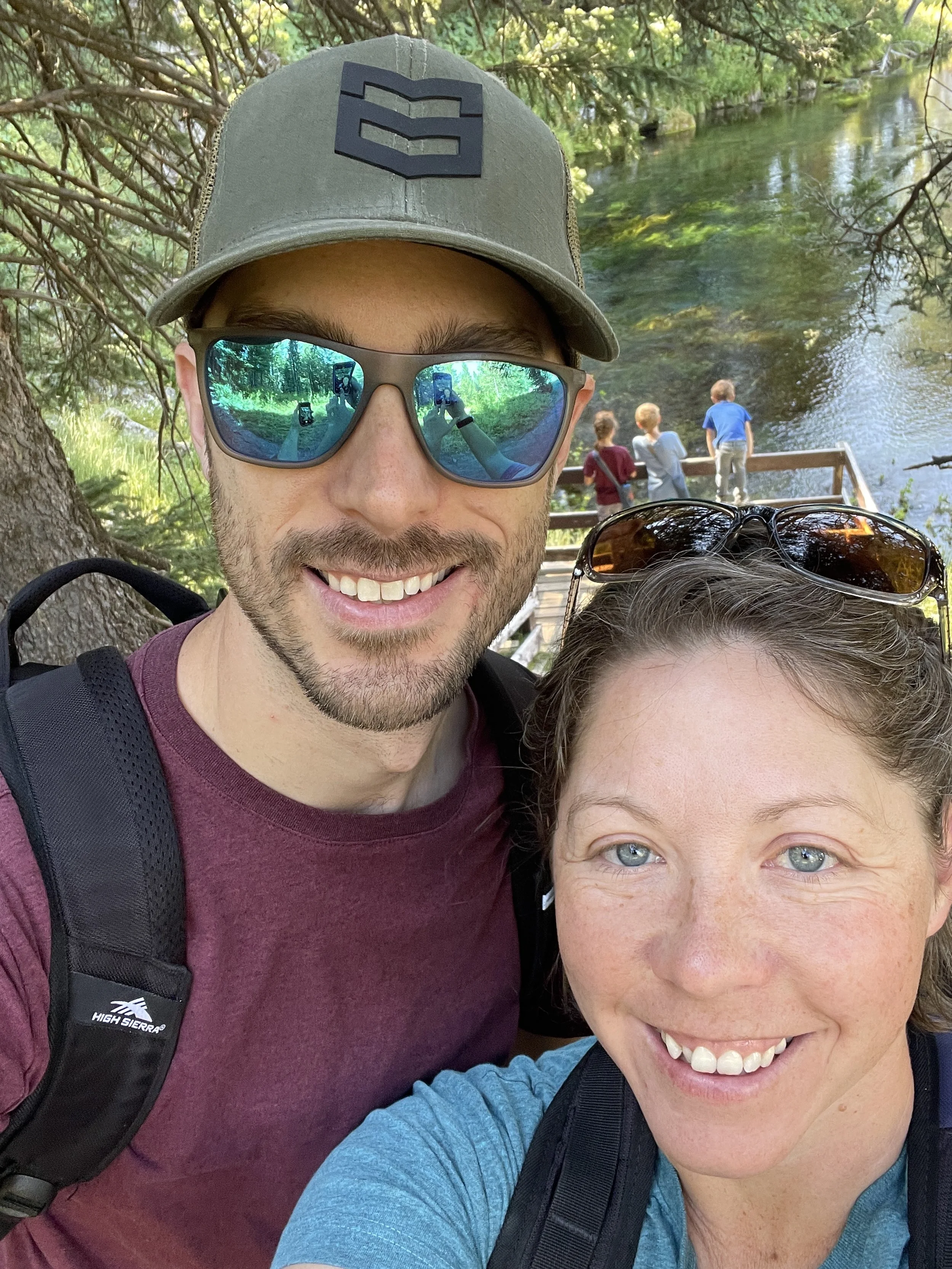 A smiling man and woman taking a selfie outdoors near a river with children playing in the background, wearing backpacks and sunglasses, under trees.