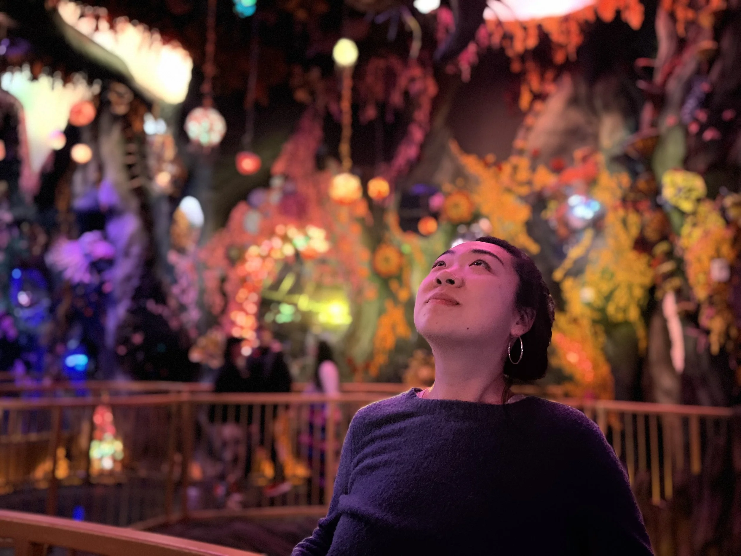 A gender expansive, queer, Asian woman looking upward in a colorful, glowing, fantasy-like environment with vibrant lights and hanging ornaments.