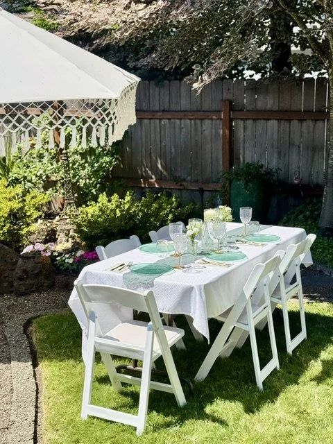 A neatly set outdoor dining table with six white chairs, glassware, and green napkins in a backyard with a wooden fence, garden plants, and a white umbrella.