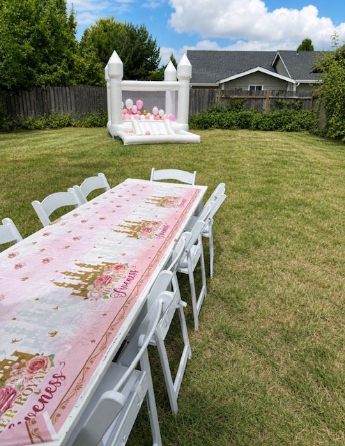 A long white outdoor table with eight white chairs around it, set up in a backyard with green grass, trees, and a wooden fence. In the background, there is a pink and white inflatable bounce house decorated with balloons.