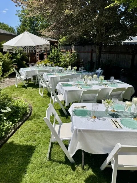 Outdoor backyard party setup with white tables and chairs, decorated with white tablecloths, floral centerpieces, and glassware, under a large white umbrella with a garden and wooden fence in the background.