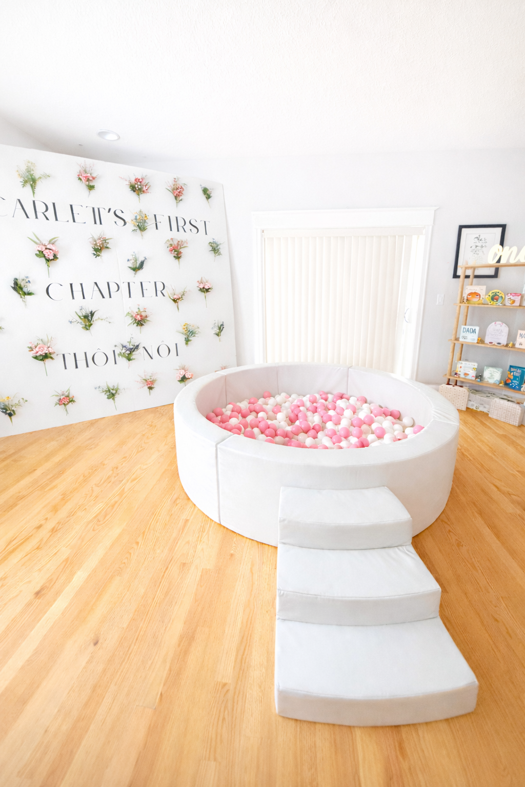 Indoor party setup with a white ball pit filled with pink and white balls, three steps leading into it, and a decorative backdrop with flowers and text in Vietnamese.