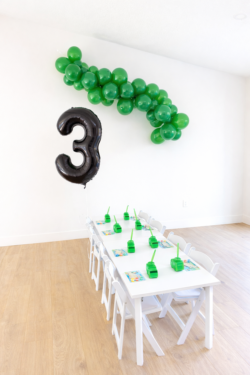 Party setup with a long white table, green juice boxes with straws, party favors, and a black number three balloon. Green balloons are arranged in a cluster on the wall, and a large black number three balloon is floating next to them.