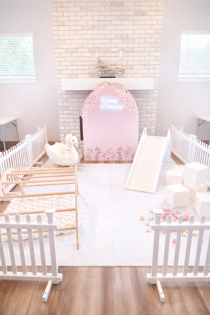 Indoor play area with white fencing, small slide, white picnic table, rocking horse, and wooden climbing arch, with decorative shelves and holiday items on the wall.