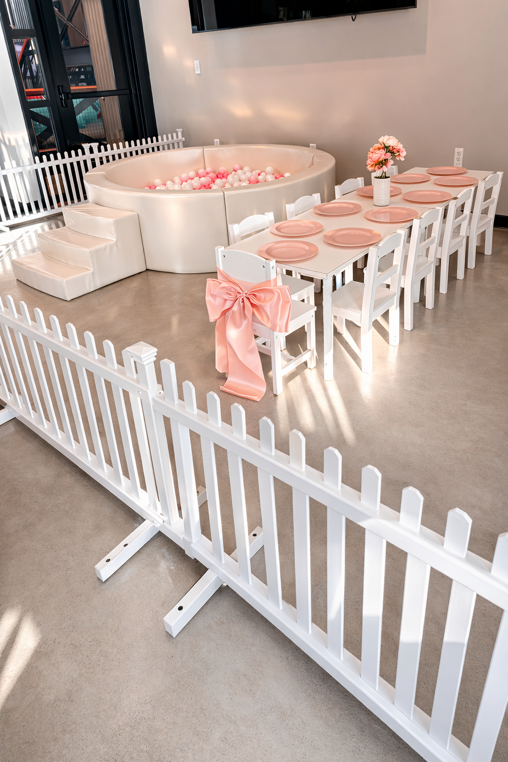 Children's party setup with a fenced area, a small white table with pink plates, a vase with pink flowers, and a ball pit filled with pink and white balls.