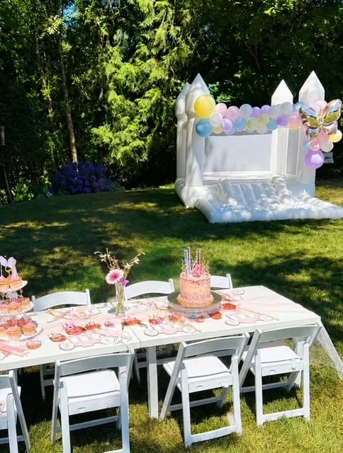 Decorated outdoor birthday party setup with a long white table, white chairs, a pink birthday cake, and an inflatable castle with balloons and butterfly decoration in a grassy yard with trees.